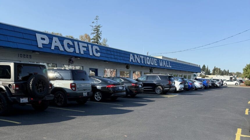 Exterior view of Pacific Antiques Mall at 10228 Pacific Avenue in Tacoma, WA, featuring large white lettering on a blue awning, a full parking lot on a sunny day, and the spacious single-story storefront with large windows.