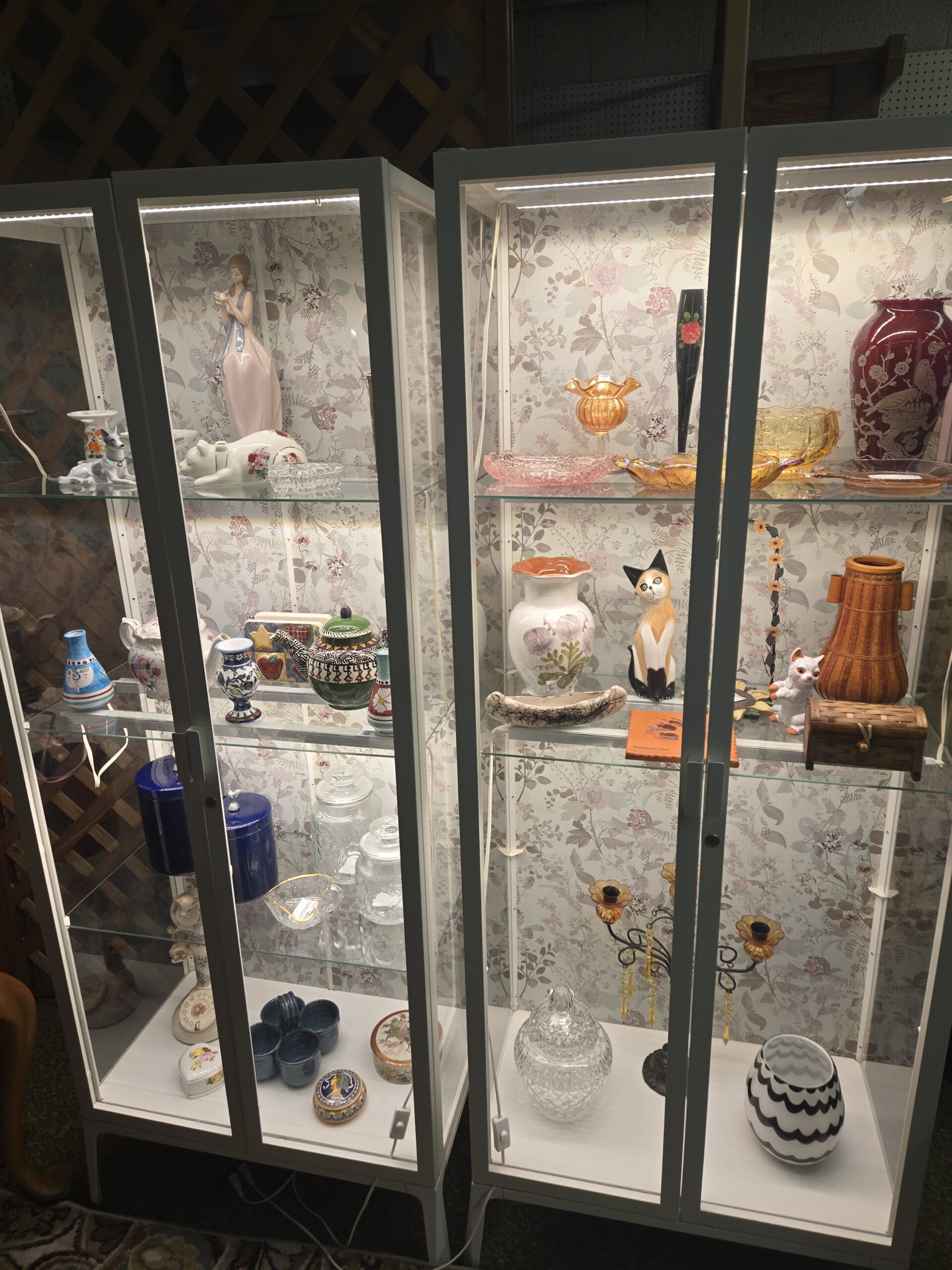 Lladro-style porcelain figurine of a young woman in pink gown, white porcelain reclining cat figurine, pink pressed glass oval dish, marigold carnival glass ruffled compote, tall black art glass bud vase with hand-painted red rose, ruby red etched glass vase with bird and floral motif, amber glass footed candy dish, white ceramic vase with purple thistle transfer print, ceramic calico cat figurine in black and white, small white porcelain seated cat figurine, amber wood-grain ceramic cylindrical vase, blue and white Dutch-style ceramic mini vase, assorted Polish pottery pieces in green and red apple motif and geometric patterns, cobalt blue ceramic canister, pressed glass apothecary jar with lid, gold-lidded milk glass trinket box with floral decal, blue ceramic egg cups, enameled trinket box with floral lid, cut crystal rose bowl, black wrought iron candelabra with amber glass flower accent, amber glass candle holders on scrollwork stand, and black and white chevron ceramic vase, at Shop 69, Pacific Antiques Mall in Tacoma, WA