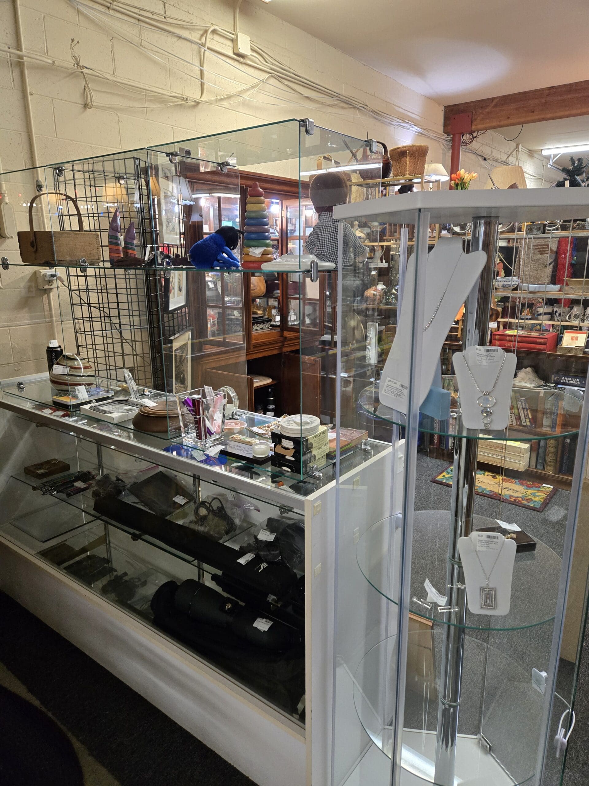 A glass and aluminum counter case at Shop 111 displaying vintage collectibles including rifle scopes and optics, small firearms-related accessories, a cobalt blue glass bear figurine, a rainbow stacking ring toy, a carved wooden head sculpture, a gingham cloth doll, and small jewelry and trinket boxes on the upper surface, alongside a tall glass tower case displaying silver-tone chain necklaces with pendant charms on white jewelry neck forms, at Shop 111, Pacific Antiques Mall in Tacoma, WA.