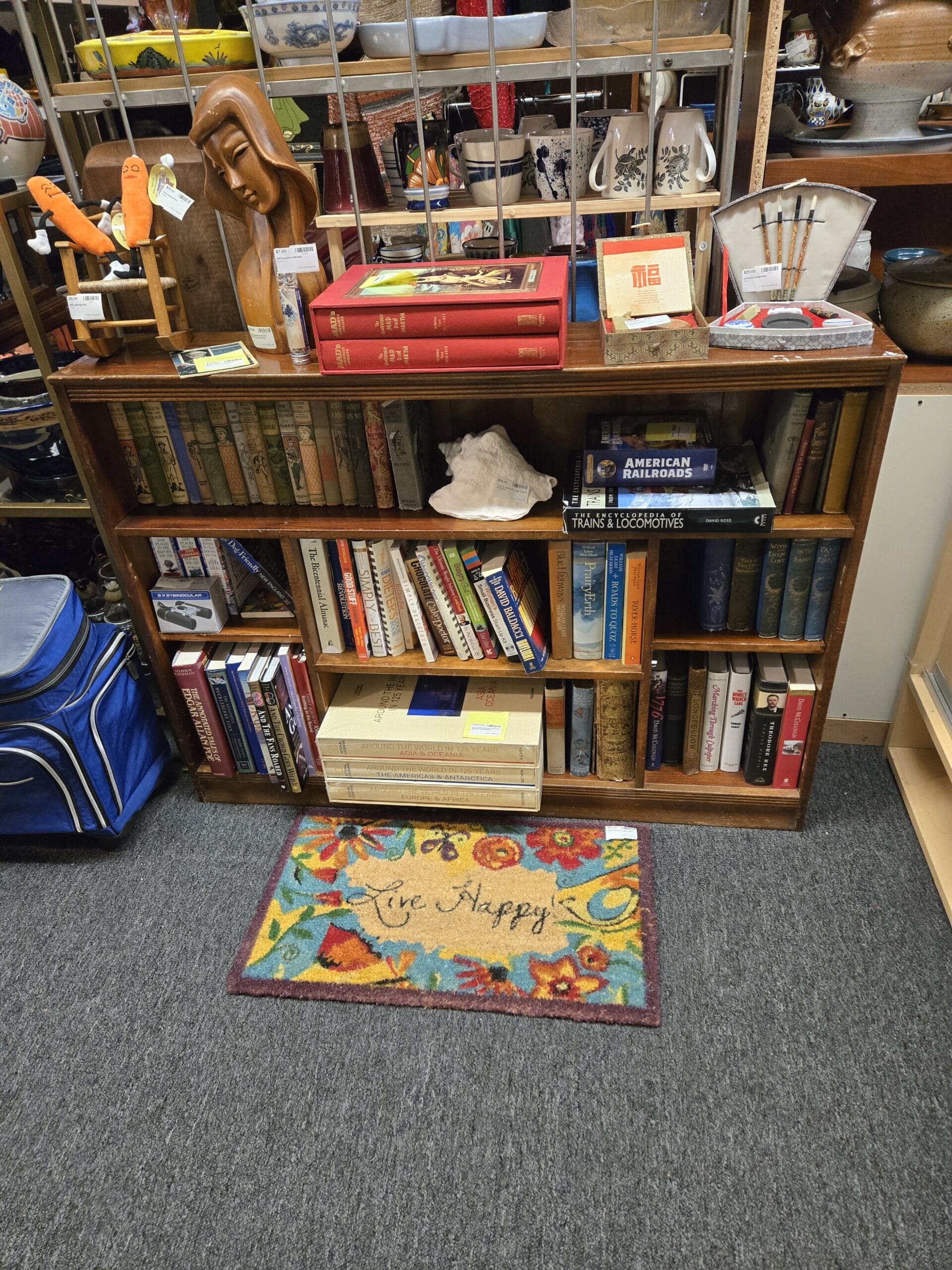 A low open oak bookcase at Shop 111 filled with a varied collection of vintage and collectible books including a set of red hardcover volumes, a copy of American Railroads, The Encyclopedia of Trains and Locomotives, Around the World in 82 Days, and additional vintage hardcover titles on history and travel, topped with a carved Art Deco wooden female bust sculpture, a large natural conch shell specimen, a Chinese red lacquer gift box, a small framed Chinese calligraphy card, and a vintage fan-shaped metal paint brush holder with bamboo brushes, with a colorful hooked accent rug reading "Live Happy" in script on the floor in front, at Shop 111, Pacific Antiques Mall in Tacoma, WA.