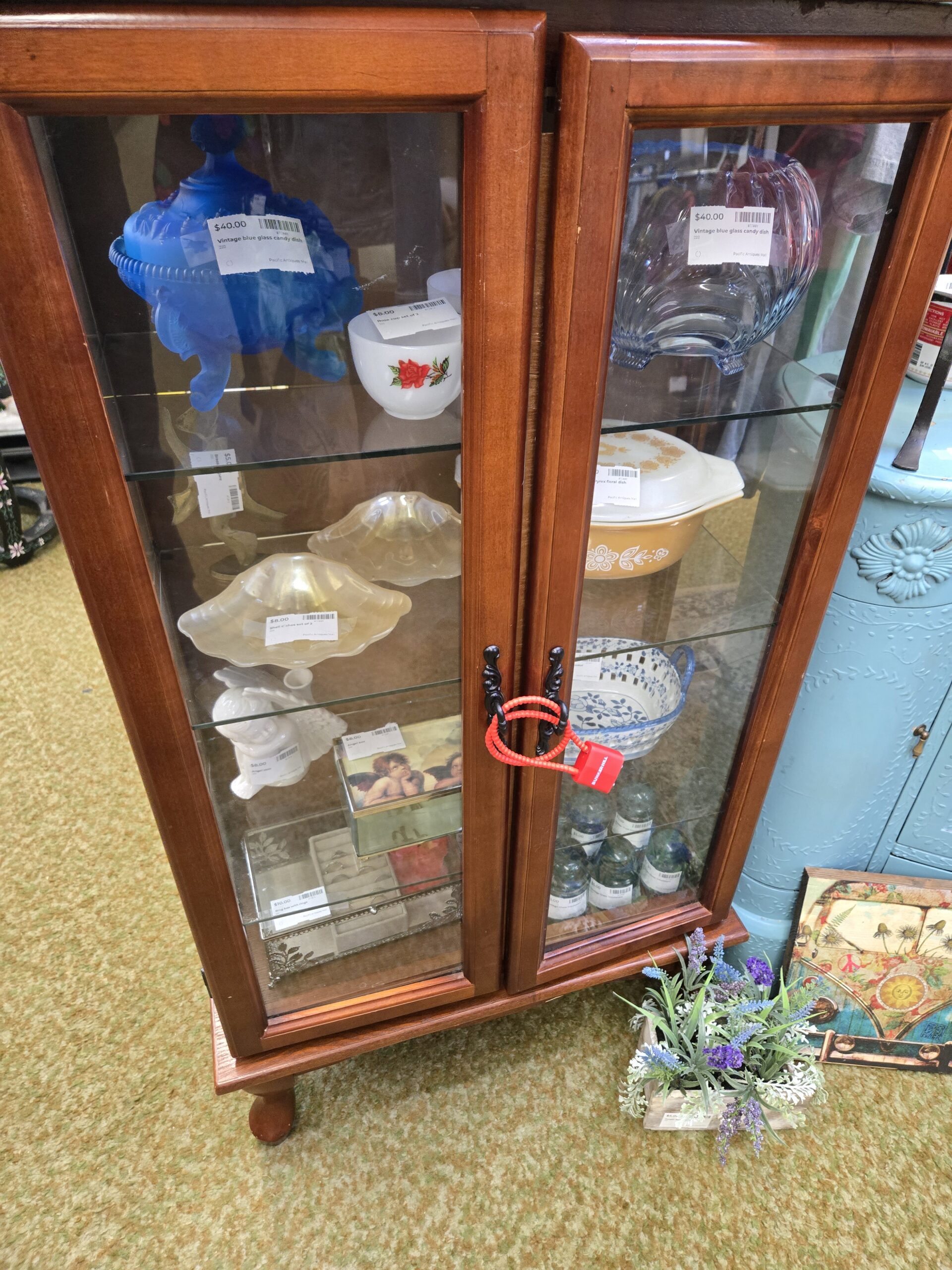 A small walnut curio cabinet with two glass panel doors on ball feet at Shop 222 displaying vintage glassware and kitchenware including a cobalt blue milk glass pedestal candy dish with ruffled rim, a clear pressed glass candy dish, a Fire-King white milk glass bowl with red rose decal, a pair of frosted white glass shell-form compote dishes on pedestal bases, a Pyrex Golden Butterfly casserole dish in amber and white, a blue and white floral transferware berry bowl, a set of small dark green glass bottles, and a set of botanical print decorative gift boxes with cherub imagery, with a faux lavender arrangement at the base, at Shop 222, Pacific Antiques Mall in Tacoma, WA.