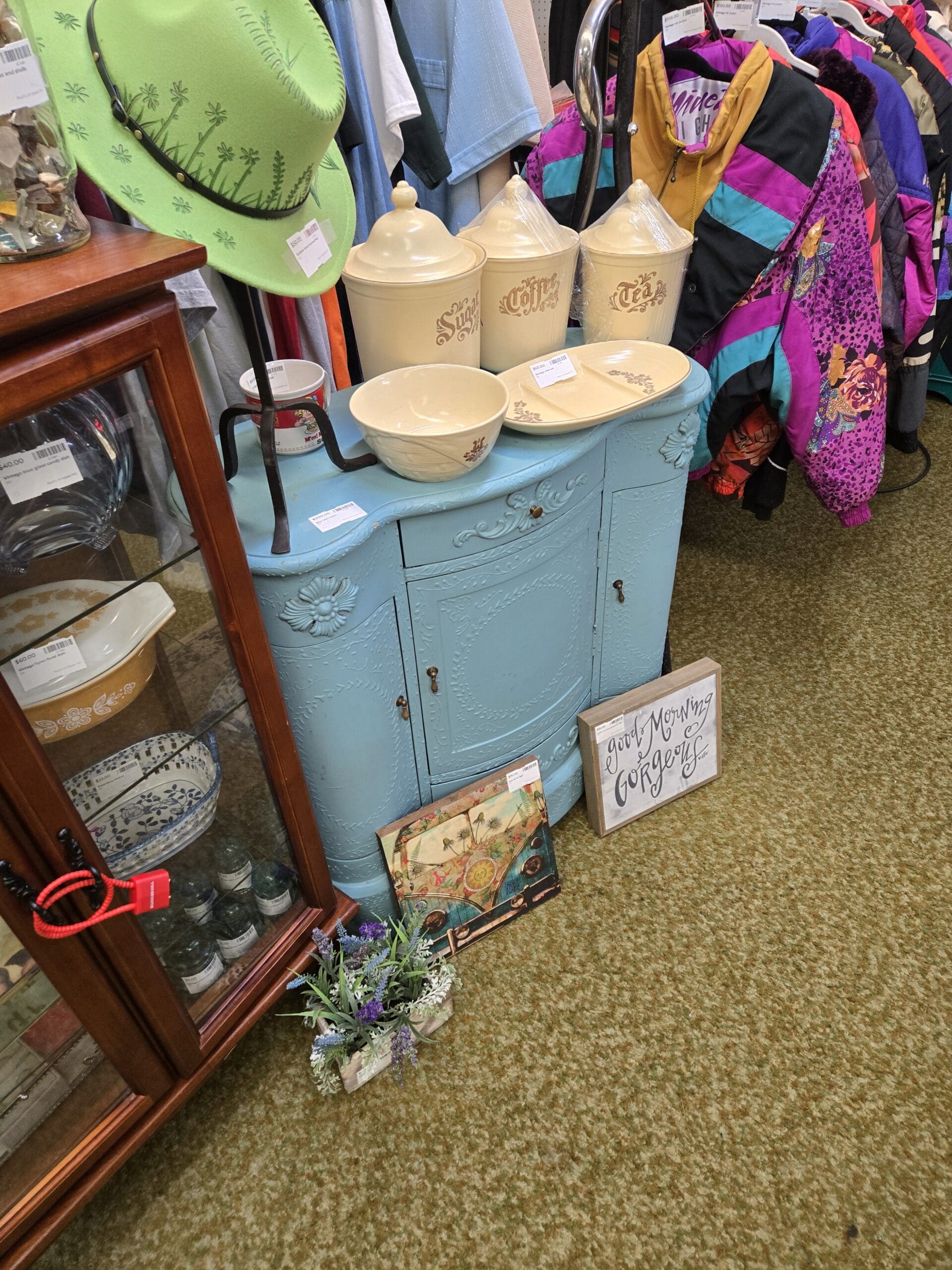A pale powder blue painted vintage carved sideboard with ornate floral and scroll relief detailing, shell medallion accents, and original bail hardware at Shop 222, topped with a set of three large cream ceramic kitchen canisters labeled Sugar, Coffee, and Tea in gold script with botanical motifs and matching lids, two white stoneware mixing bowls, and a black metal mug tree rack, displayed alongside a "Good Morning Gorgeous" wood sign, a colorful vintage Volkswagen bus print, and a faux lavender plant, with a green embroidered wide-brim felt hat and glass curio cabinet visible to the left and colorful vintage ski jackets on a rack to the right, at Shop 222, Pacific Antiques Mall in Tacoma, WA.