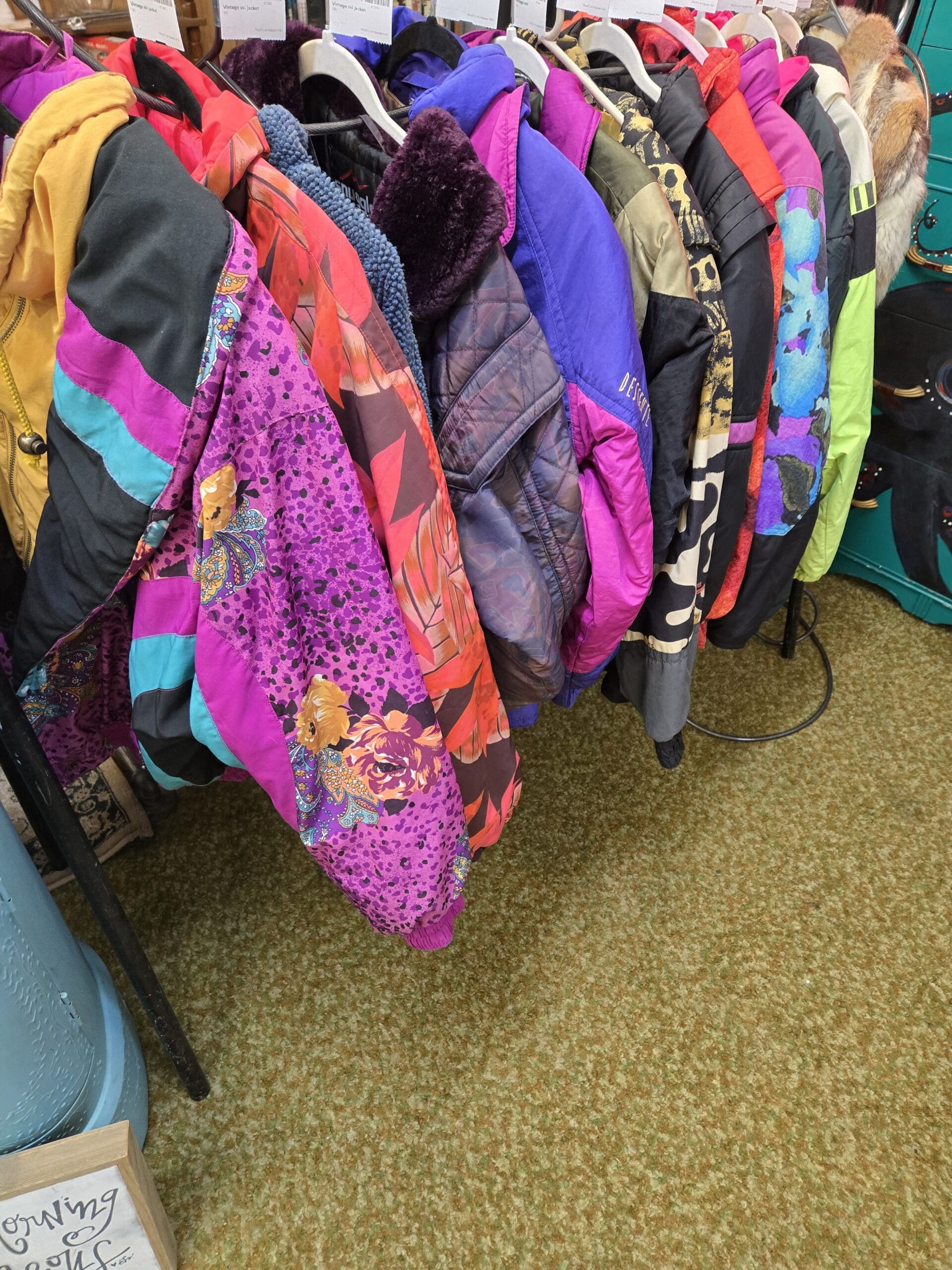 A densely packed vintage ski jacket and outerwear clothing rack at Shop 222 displaying a vivid collection of 1980s and 1990s colorblock and printed ski jackets including a purple and black leopard spot print jacket with teal and magenta colorblock panels, a red and orange abstract tropical leaf print jacket, a purple quilted puffer with faux fur collar, a Descente branded purple and gray ski jacket, a black jacket with blue floral print, a leopard print jacket with gold and red colorblock accents, and additional jackets in bright yellow, neon green, olive, and multicolor abstract prints, at Shop 222, Pacific Antiques Mall in Tacoma, WA.