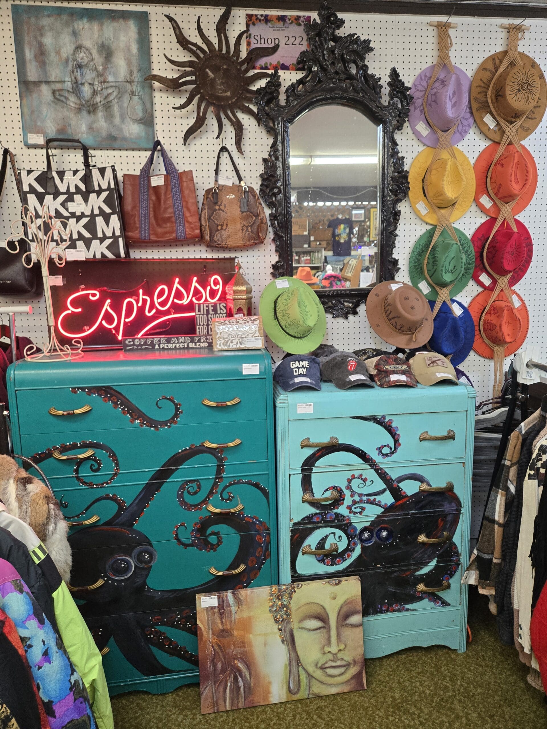 A pegboard wall display at Shop 222 featuring two teal painted dressers with elaborate hand-painted black octopus tentacle artwork with gold drawer pulls, a black ornate Baroque-style wall mirror with carved acanthus leaf frame, a black metal sunburst wall sculpture, a red neon Espresso sign with coffee-themed text, Michael Kors logo canvas tote bag, brown leather Coach handbag, brown snake-print structured handbag, a rainbow wall display of wide-brim felt and suede hats in purple, tan, green, yellow, orange, red, and coral tones, a collection of baseball caps including a Game Day cap and Rolling Stones logo cap, and a hand-painted Buddha portrait canvas at the base, at Shop 222, Pacific Antiques Mall in Tacoma, WA.