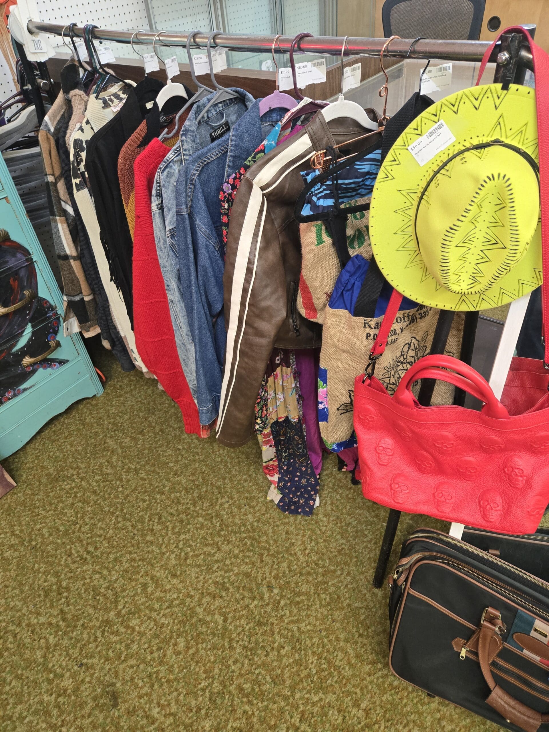 A clothing rack at Shop 222 displaying a mix of vintage women's tops, jackets, and accessories including a black and white cowhide print knit top, a red chunky knit sweater, a Thrill brand denim jacket, a brown leather bomber jacket with white racing stripe, a floral print blouse, colorful scarves, a burlap Kauai coffee estate market tote bag, a yellow neon wide-brim felt hat with black sunburst stitching, a red embossed skull motif leather handbag, and a black and brown travel duffel bag on the floor, at Shop 222, Pacific Antiques Mall in Tacoma, WA.