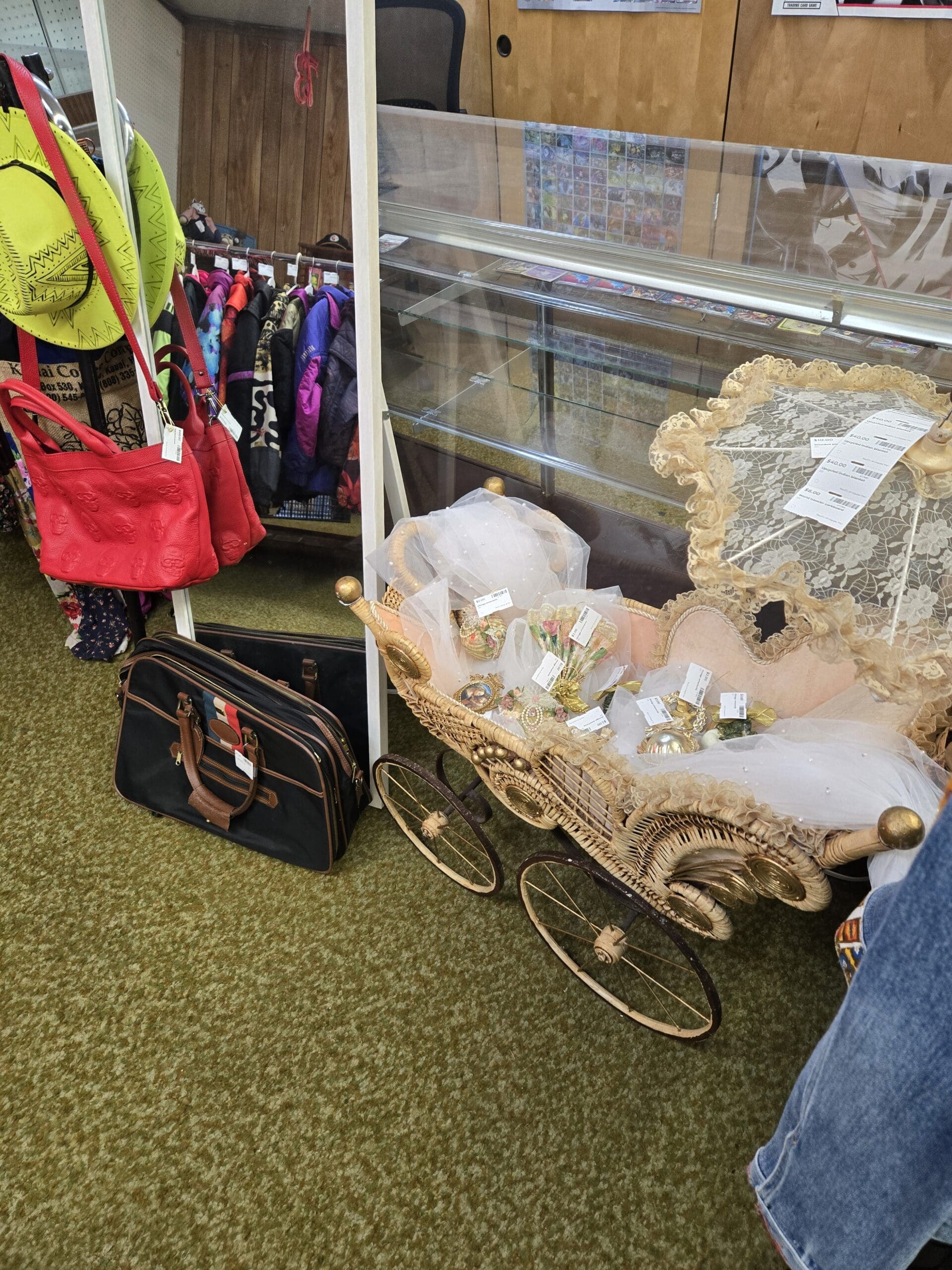A vintage wicker and gilt metal antique doll pram baby carriage at Shop 222 with a cream lace canopy hood, pink satin interior, spoked metal wheels, and brass ball finials, filled with packaged vintage floral brooch and jewelry items, displayed on the shop floor beside a black and brown striped travel duffel bag, with a red embossed skull handbag and yellow wide-brim hat hanging on a rack to the left and a trading card display case visible in the background, at Shop 222, Pacific Antiques Mall in Tacoma, WA.