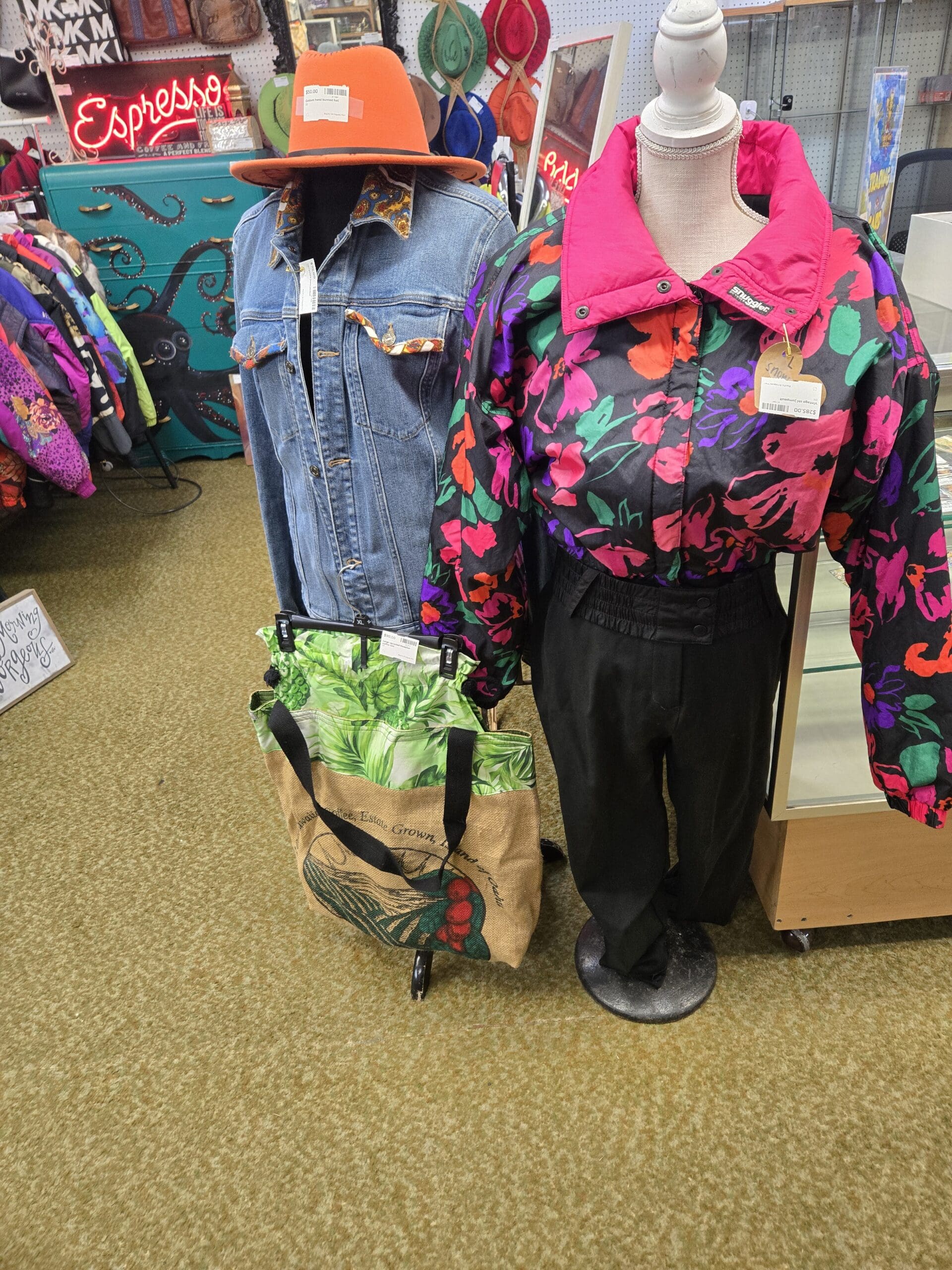Two mannequin torsos at Shop 222 displaying vintage clothing including a medium blue wash denim trucker jacket with a paisley print bandana knotted in the chest pocket and an orange wide-brim felt hat, and a Snuggler brand vintage 1980s cropped ski jacket in black with bold hot pink, orange, green, and purple abstract floral print and a fuchsia pink collar, paired with black high-waist trousers and a rhinestone chain necklace, with a burlap and green tropical print market tote bag displayed on a rack between them, and the octopus dressers, neon Espresso sign, and colorful vintage jacket rack visible in the background, at Shop 222, Pacific Antiques Mall in Tacoma, WA.