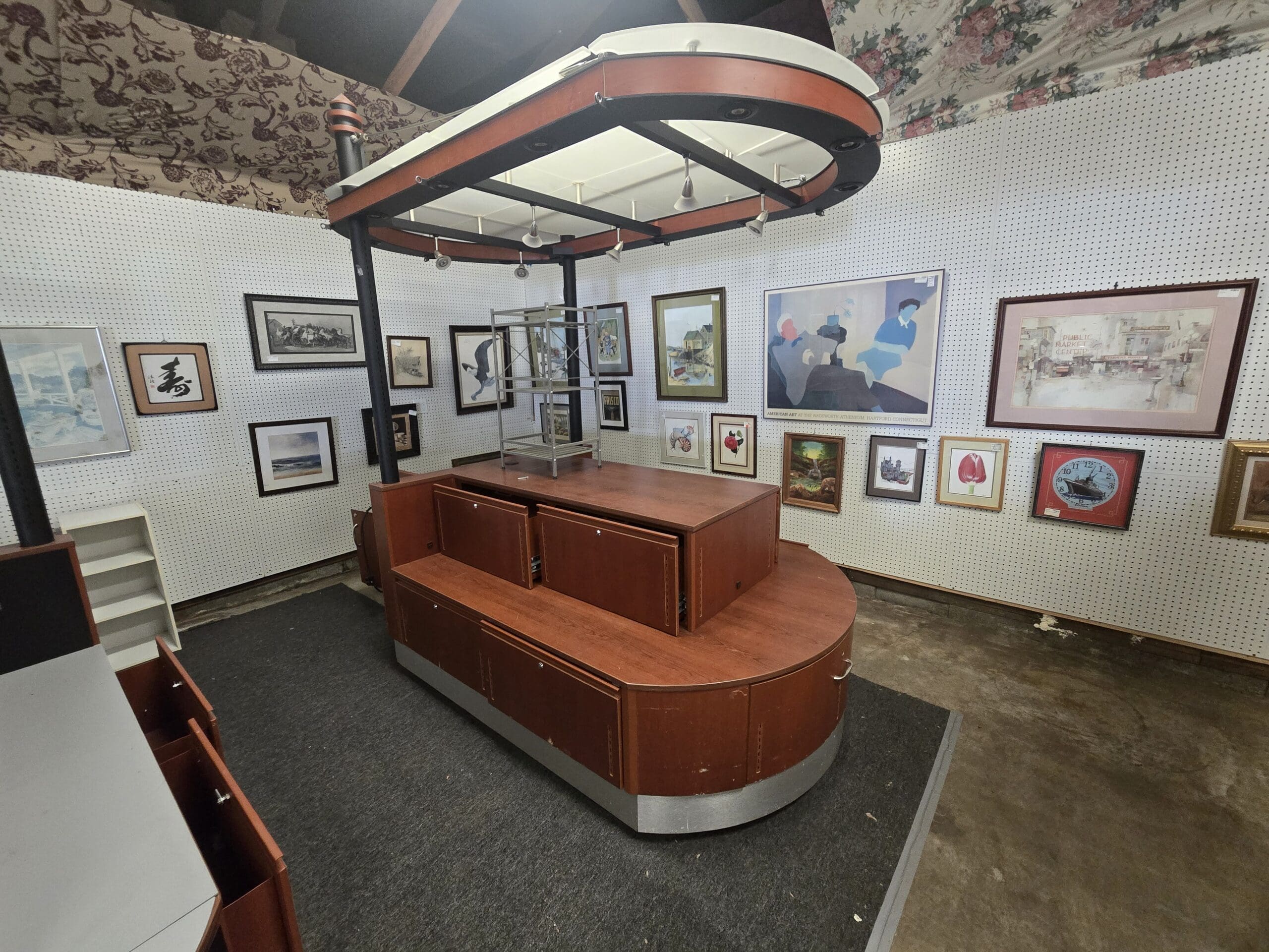 A single oval cherry wood laminate retail kiosk island at West Wing Wares with a two-tiered stepped counter, locking lower cabinet storage, and overhead canopy lighting rig with track spotlights.