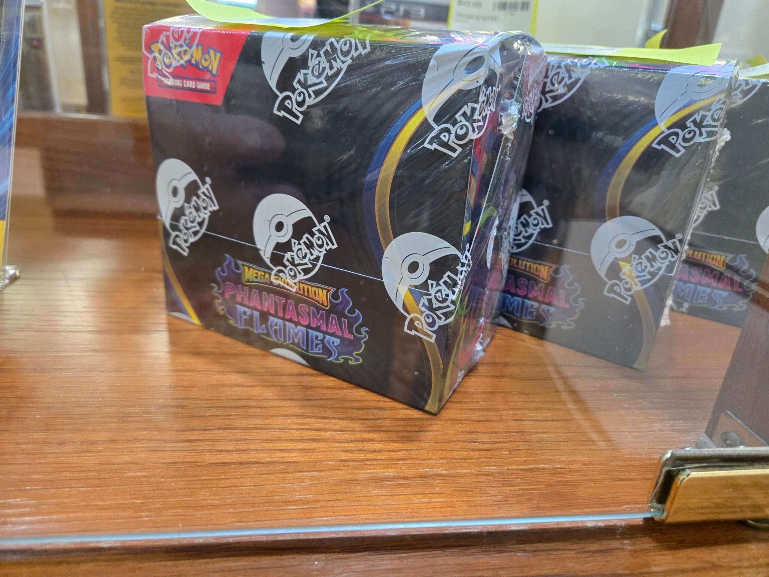 Sealed Pokemon Trading Card Game Mega Evolution Phantasmal Flames booster boxes in black packaging with repeated white Poke Ball logos, resting on a wood shelf inside a glass cabinet at Shop 117, Pacific Antiques Mall in Tacoma, WA.