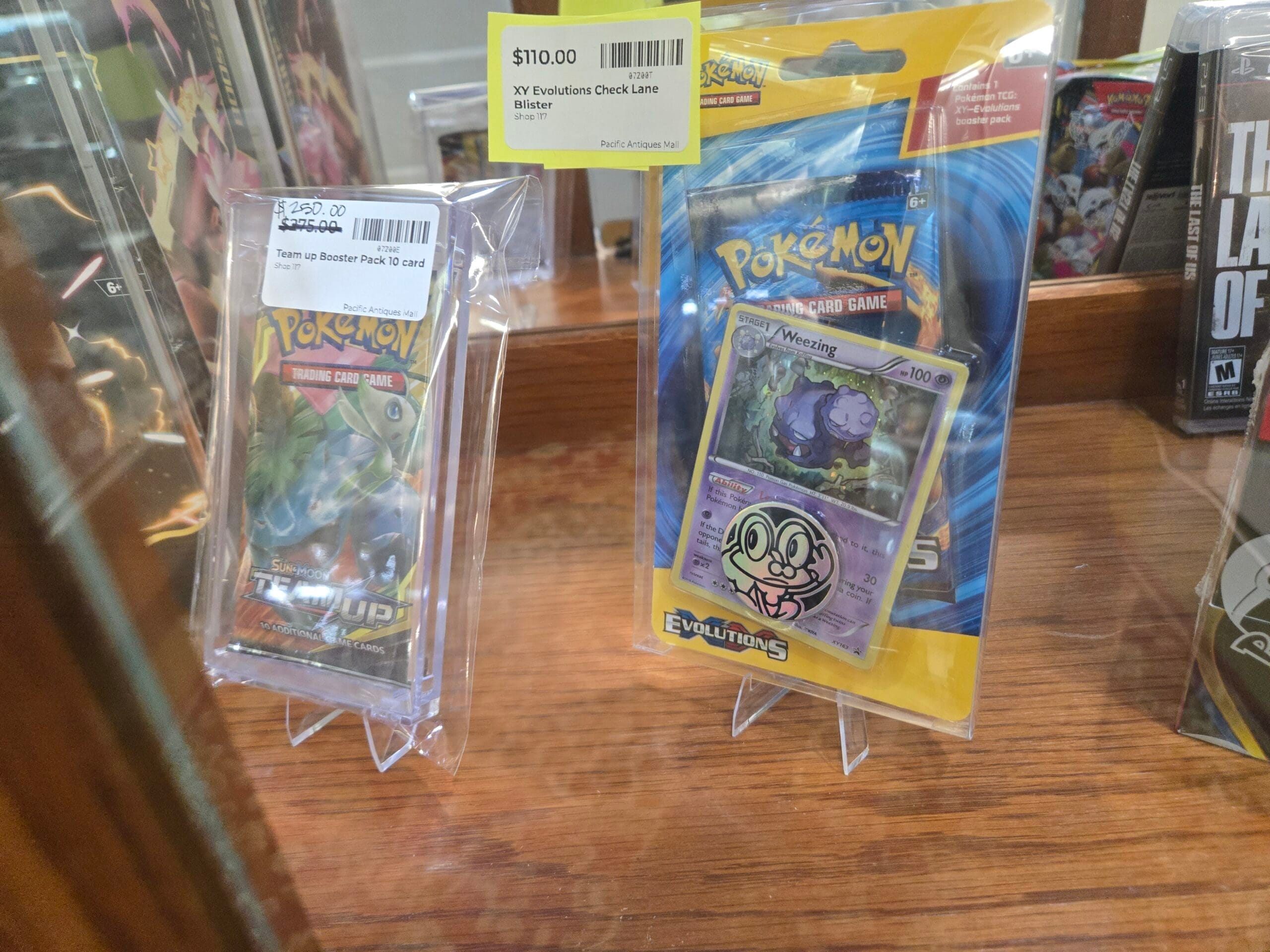 A sealed Pokemon Trading Card Game Sun and Moon Team Up booster pack and a Pokemon XY Evolutions check-lane blister pack showing a Weezing card, both upright on clear easels beside a sealed The Last of Us PlayStation 3 game at Shop 117, Pacific Antiques Mall in Tacoma, WA.