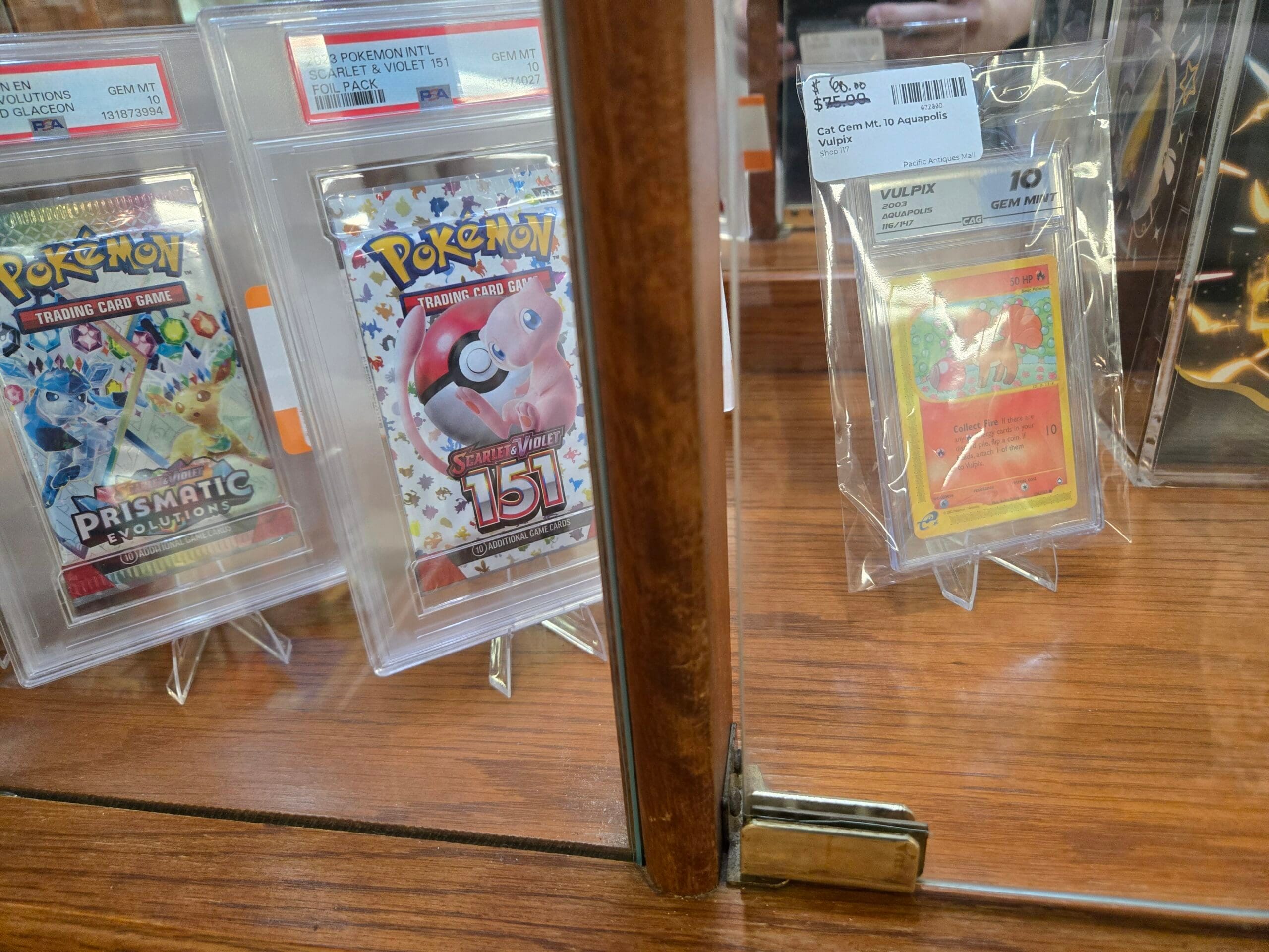 Graded and sealed Pokemon cards on clear stands, including a PSA Gem Mint 10 Prismatic Evolutions Glaceon booster pack, a PSA Gem Mint 10 2023 Scarlet and Violet 151 Mew foil pack, and a CGC Gem Mint 10 2003 Aquapolis Vulpix card at Shop 117, Pacific Antiques Mall in Tacoma, WA.