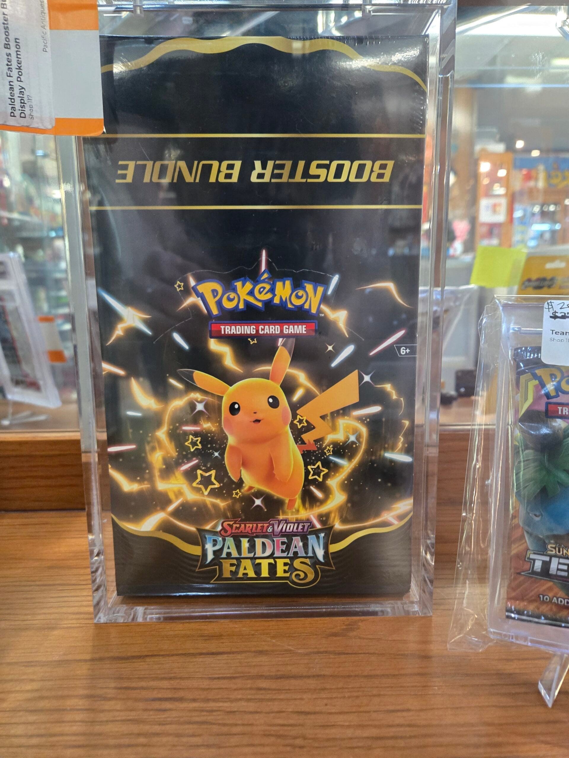 A sealed Pokemon Trading Card Game Scarlet and Violet Paldean Fates booster bundle in black and gold packaging with shiny Pikachu artwork, held in a clear acrylic case on a wood shelf at Shop 117, Pacific Antiques Mall in Tacoma, WA.