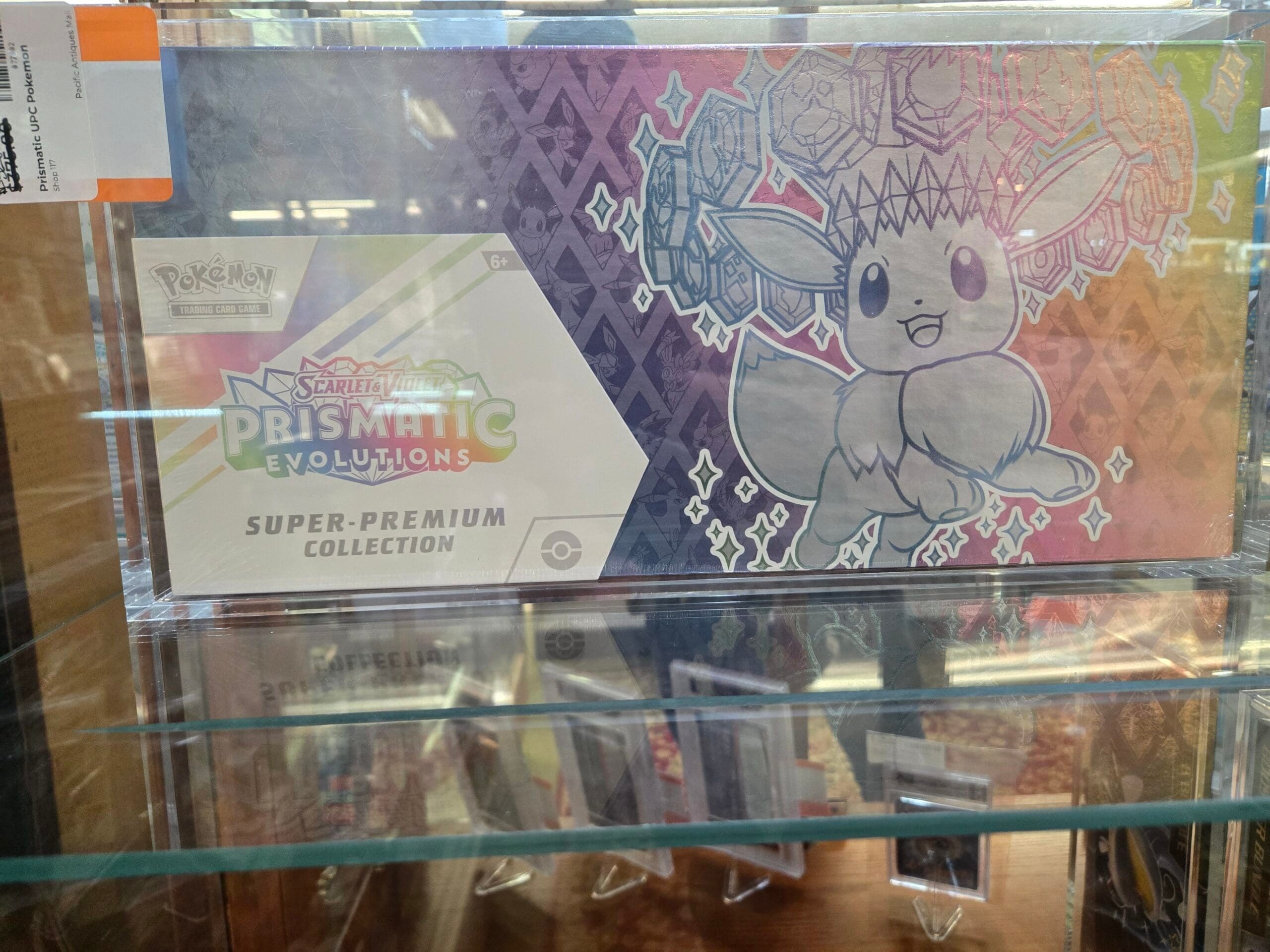 A sealed Pokemon Trading Card Game Scarlet and Violet Prismatic Evolutions Super-Premium Collection box with holographic Eevee artwork, displayed above rows of graded card slabs on clear stands inside a glass cabinet at Shop 117, Pacific Antiques Mall in Tacoma, WA.