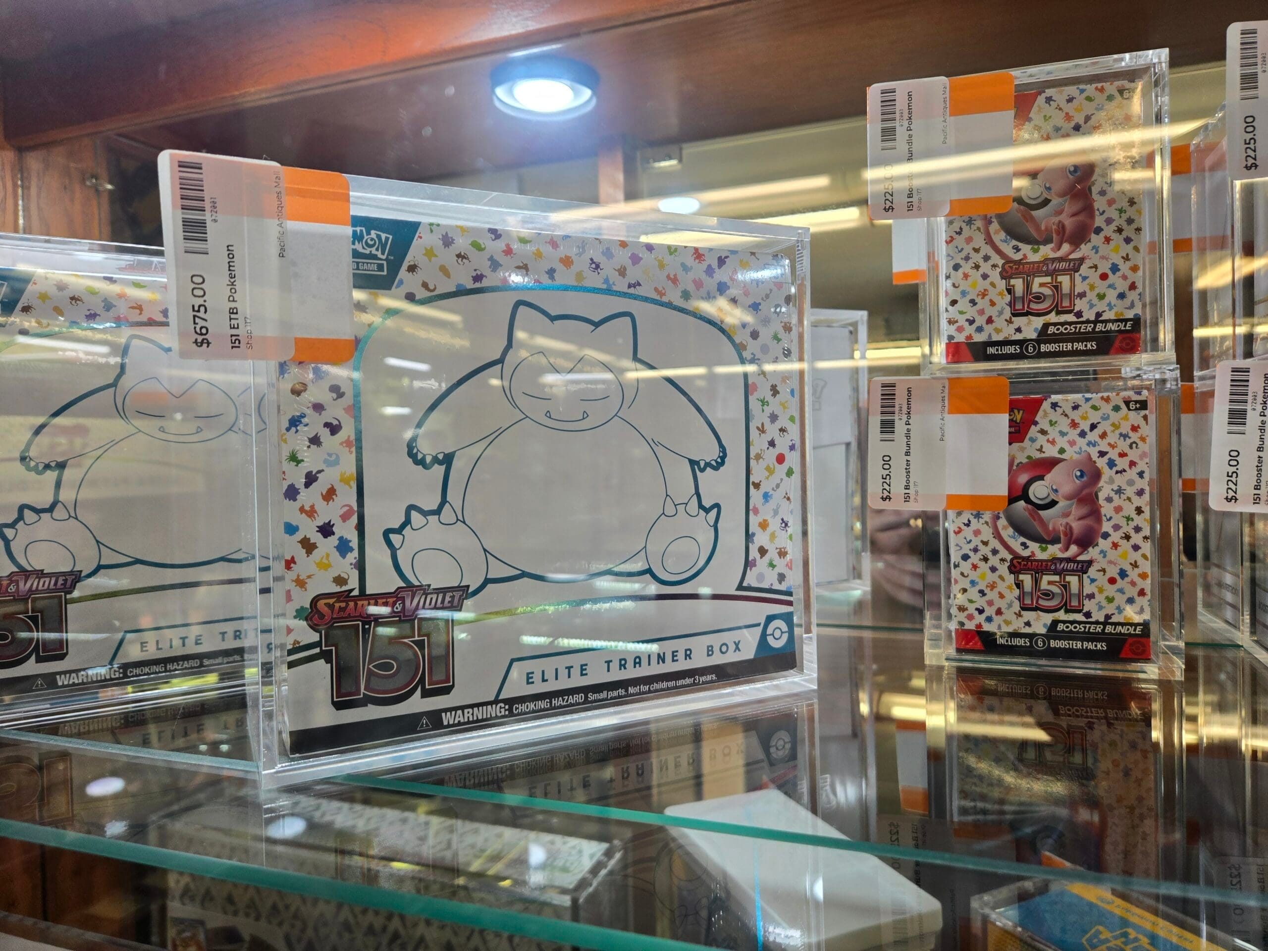 A sealed Pokemon Trading Card Game Scarlet and Violet 151 Elite Trainer Box with Snorlax line-art on a white box, beside two Scarlet and Violet 151 booster bundles featuring Mew on a Poke Ball, each labeled six booster packs and held in clear acrylic cases at Shop 117, Pacific Antiques Mall in Tacoma, WA.