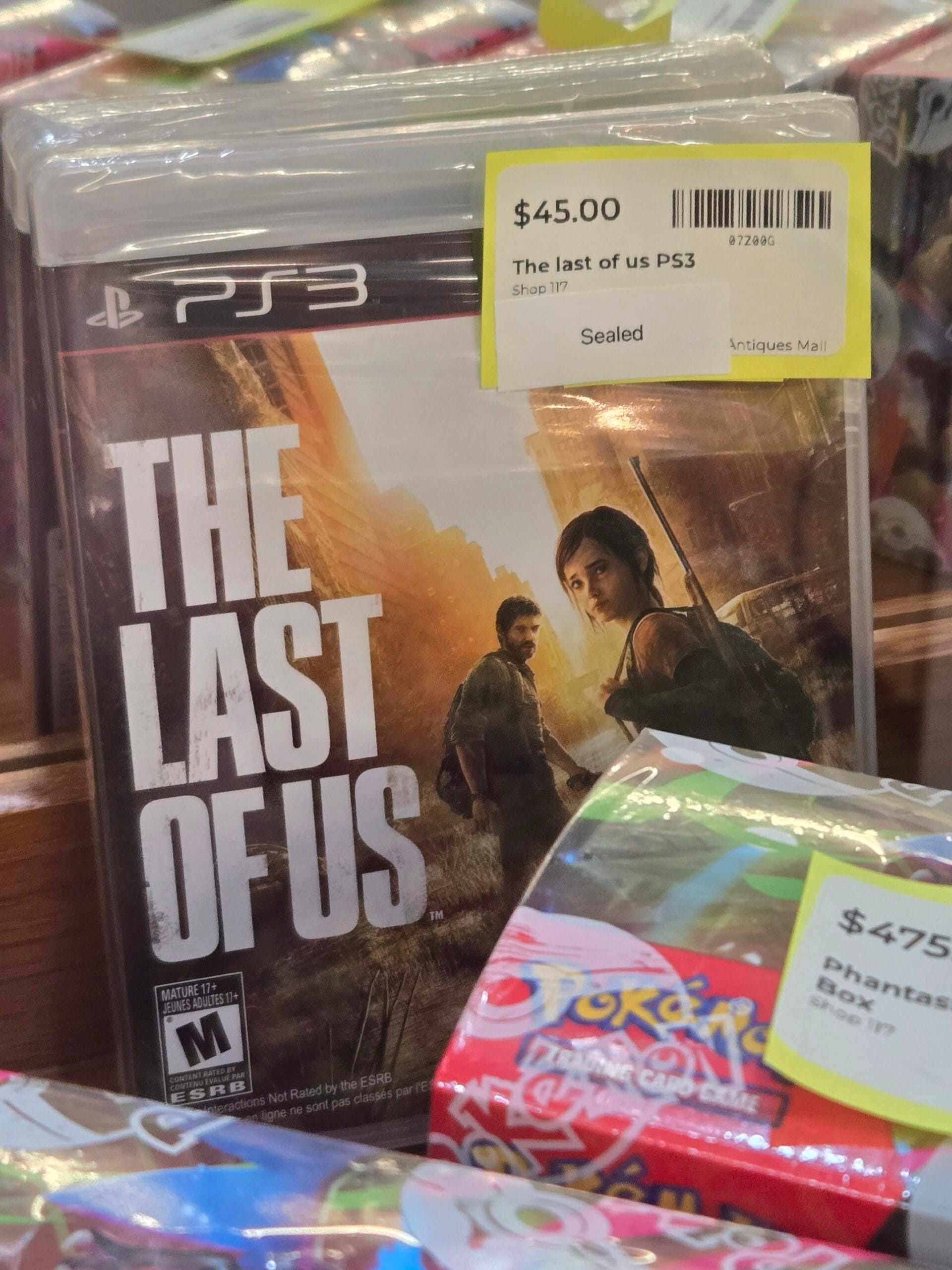 A factory-sealed The Last of Us game for PlayStation 3 in its original case, beside a red and black Pokemon Mega Evolution Phantasmal Flames box at Shop 117, Pacific Antiques Mall in Tacoma, WA.