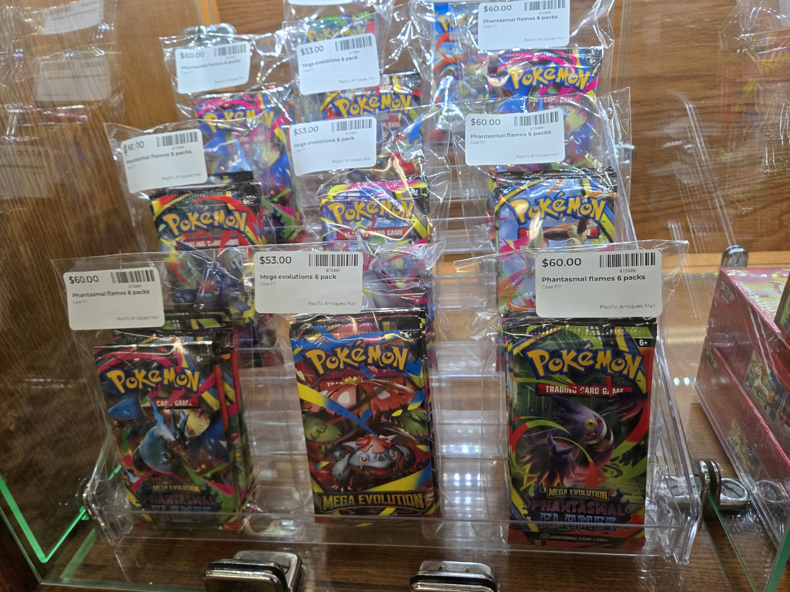 Rows of sealed Pokemon Trading Card Game booster-pack bundles on clear risers, including Mega Evolution Phantasmal Flames six-packs with blue and black Charizard artwork and Mega Evolution six-packs with green Venusaur artwork at Shop 117, Pacific Antiques Mall in Tacoma, WA.