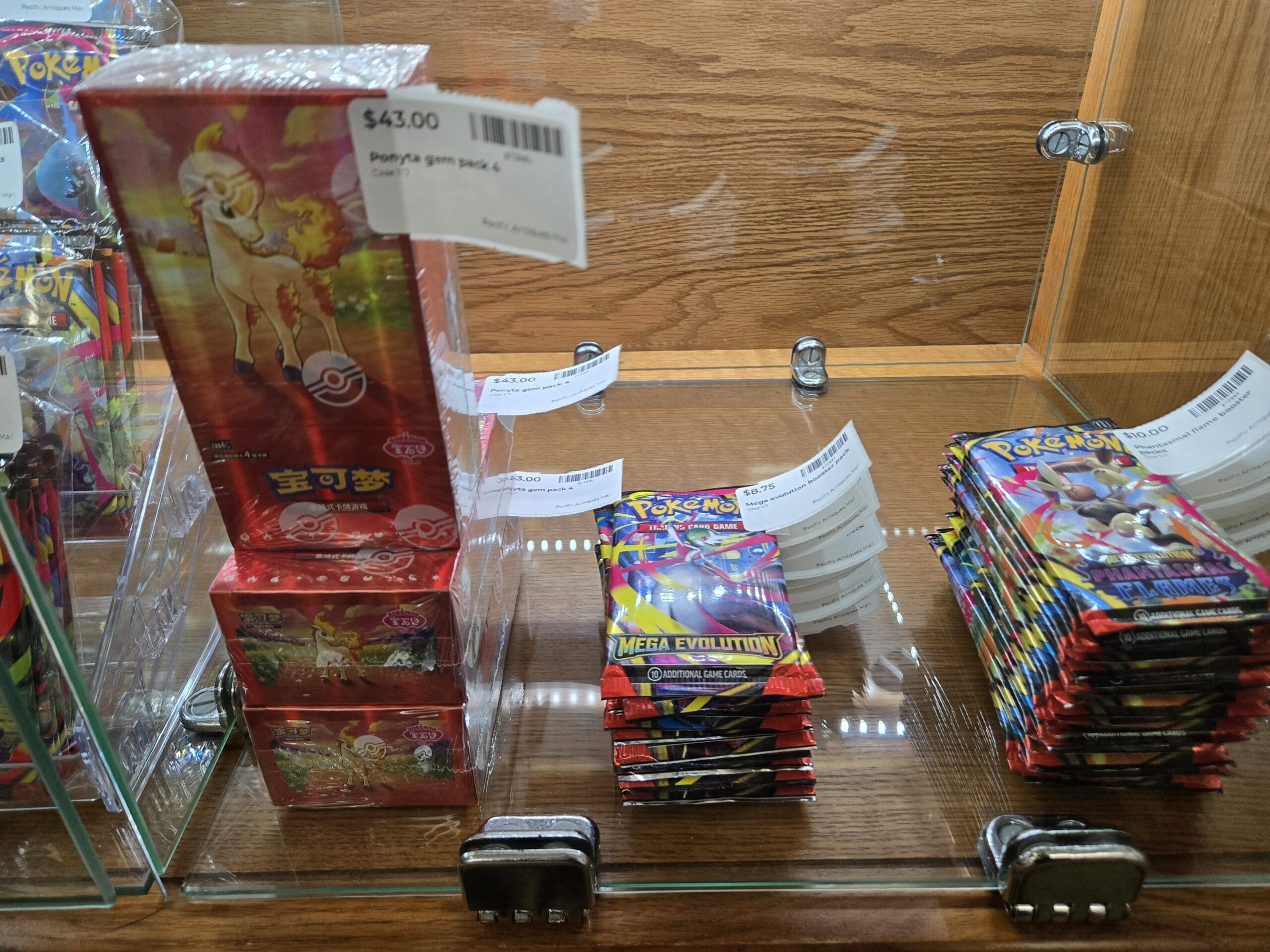 A stack of Chinese-language Pokemon Trading Card Game boxes featuring Ponyta in red packaging, beside bundled loose Mega Evolution booster packs and Mega Evolution Phantasmal Flames booster packs inside a glass cabinet at Shop 117, Pacific Antiques Mall in Tacoma, WA.