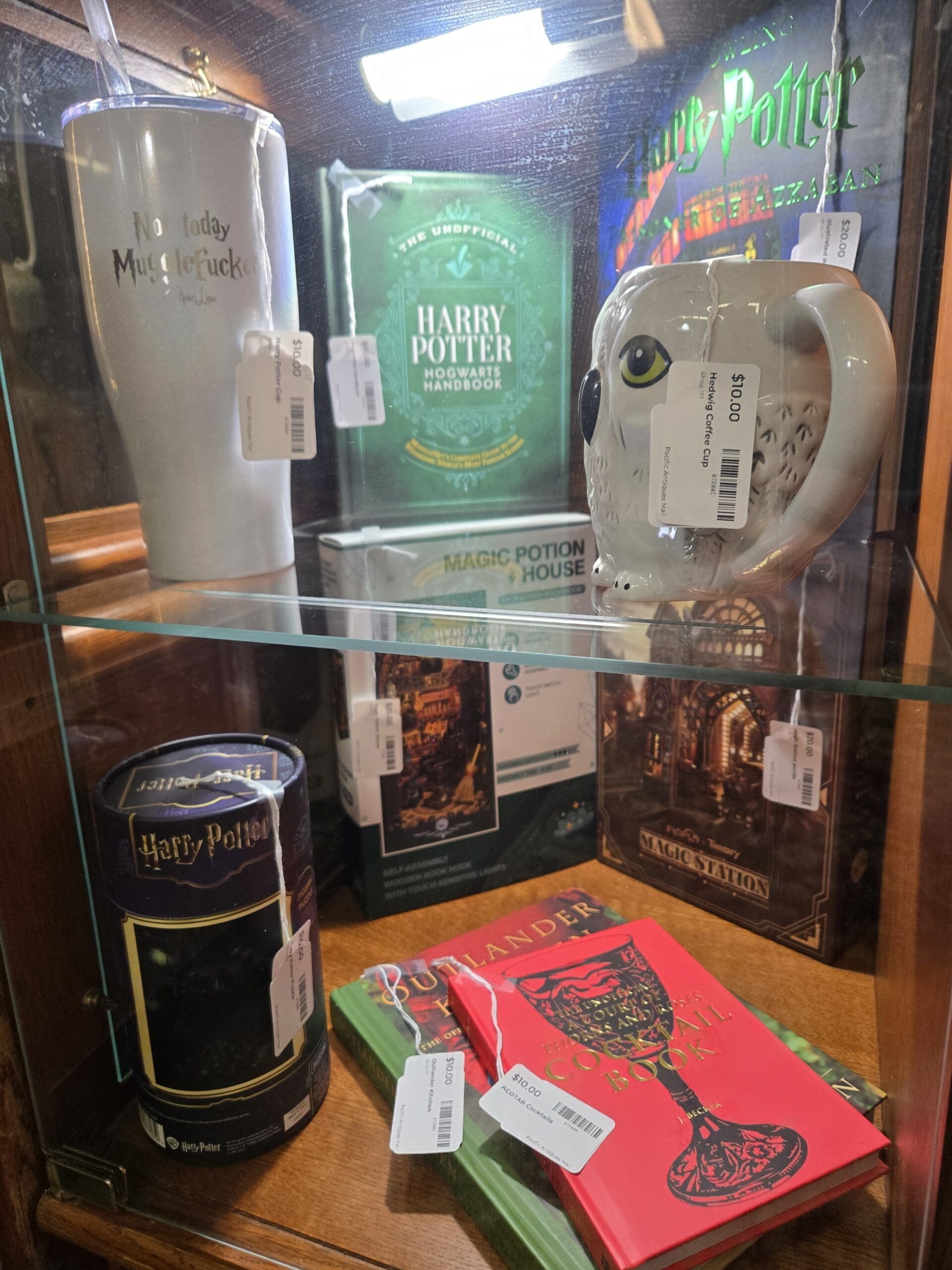 Pop-culture collectibles including a frosted Hedwig owl-shaped glass mug, an illustrated Harry Potter and the Prisoner of Azkaban edition, The Unofficial Harry Potter Hogwarts Handbook, a Magic Potion House title, a Magic Station box, a frosted Harry Potter novelty tumbler, a black Harry Potter tin, and the red Official Outlander Kitchen cookbook at Shop 117, Pacific Antiques Mall in Tacoma, WA.