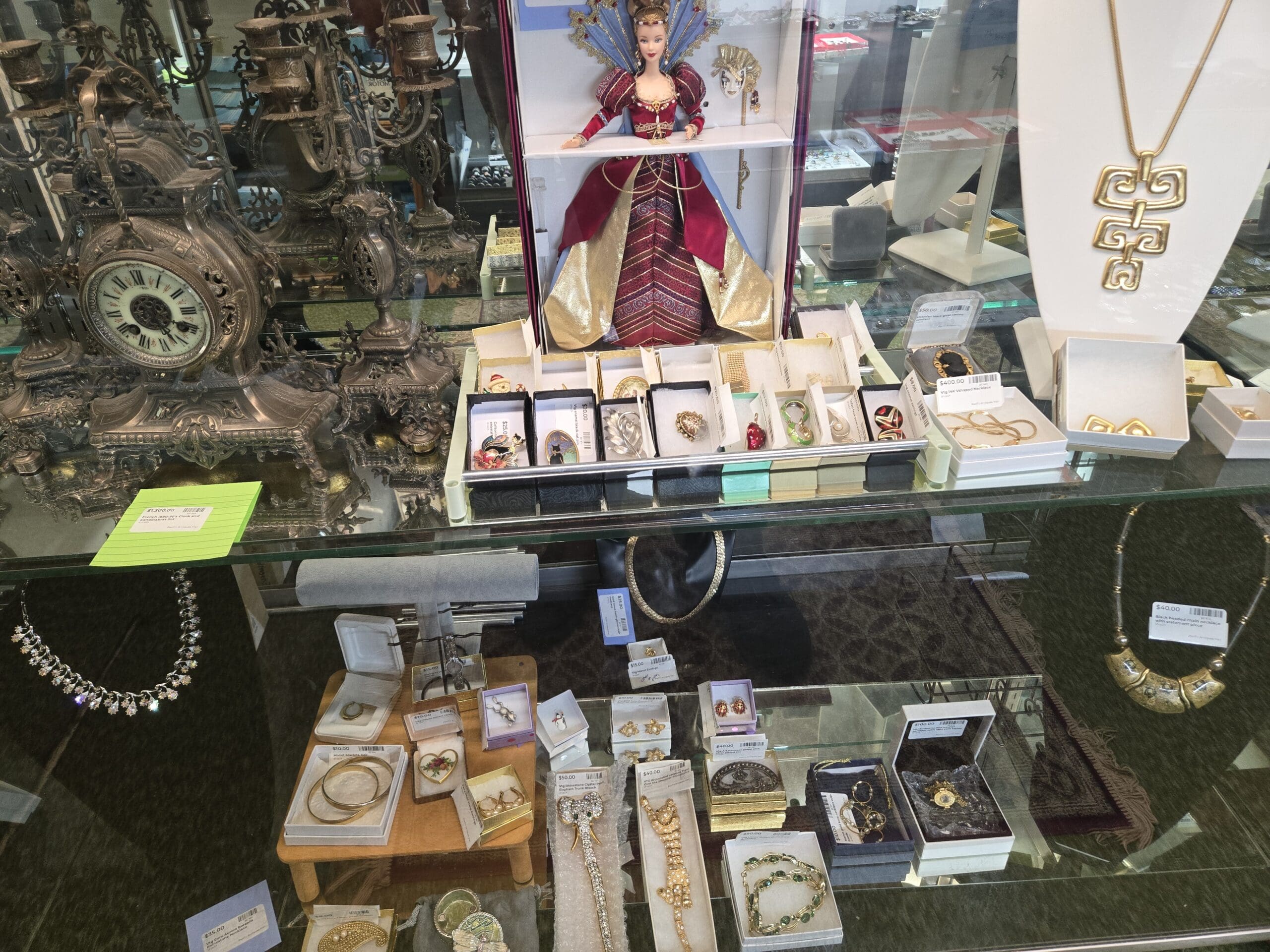Jewelry case interior with an ornate silver-tone candelabra and matching mantel clock with a Roman numeral dial, a Venetian Opulence collector Barbie doll in original box wearing red and gold robes with a peacock-feather mask, a gold modernist pendant necklace on a white bust, vintage brooches in white boxes including rose, heart, and cat motifs, a rhinestone necklace, a gold-tone bib necklace with green stones, pearl and rhinestone earrings, and bangle bracelets on a wood riser at Shop 24, Pacific Antiques Mall in Tacoma, WA.