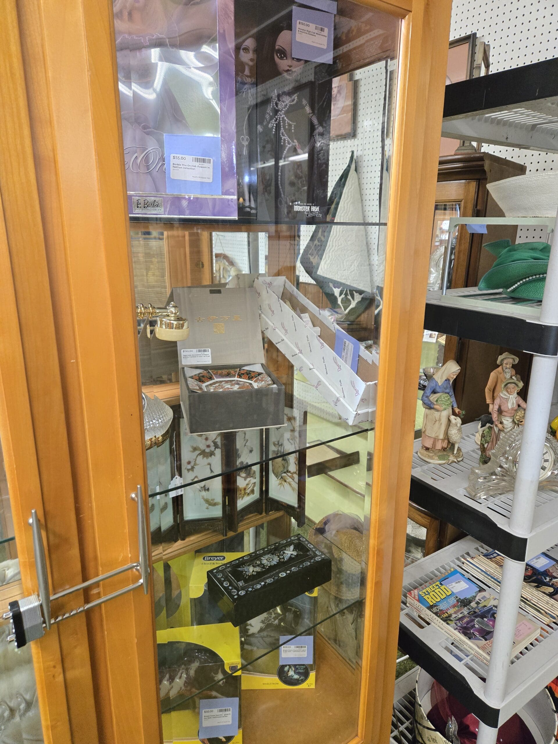 Cabinet interior showing a Monster High doll in original box, a Barbie collector doll in purple and silver packaging, a small Imari-style polychrome porcelain bowl in its presentation box on a painted Asian-style folding screen with bird motifs, a black lacquer mother-of-pearl inlaid dragon box, and a Breyer horse box on the bottom shelf, with a side shelf of vintage straw hats, bisque figurines of an elderly couple in folk dress, and a stack of Street Rod magazines at Shop 24, Pacific Antiques Mall in Tacoma, WA.