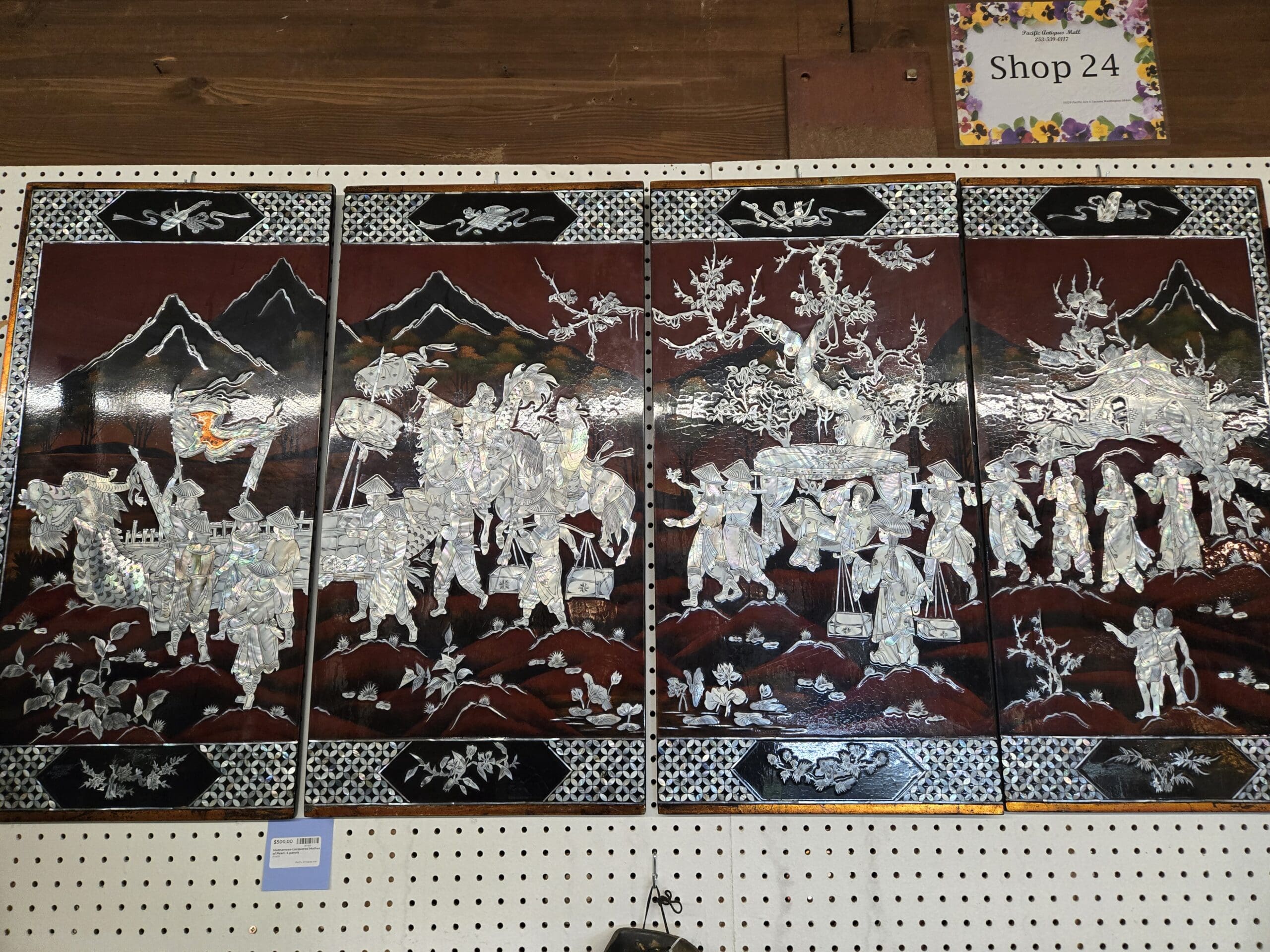 Four black lacquer wall panels with elaborate mother-of-pearl inlay depicting mountain landscapes, village figures with palanquins and animals, plum blossom trees, and waterside scenes, hung as a set under the Shop 24 sign at Shop 24, Pacific Antiques Mall in Tacoma, WA.