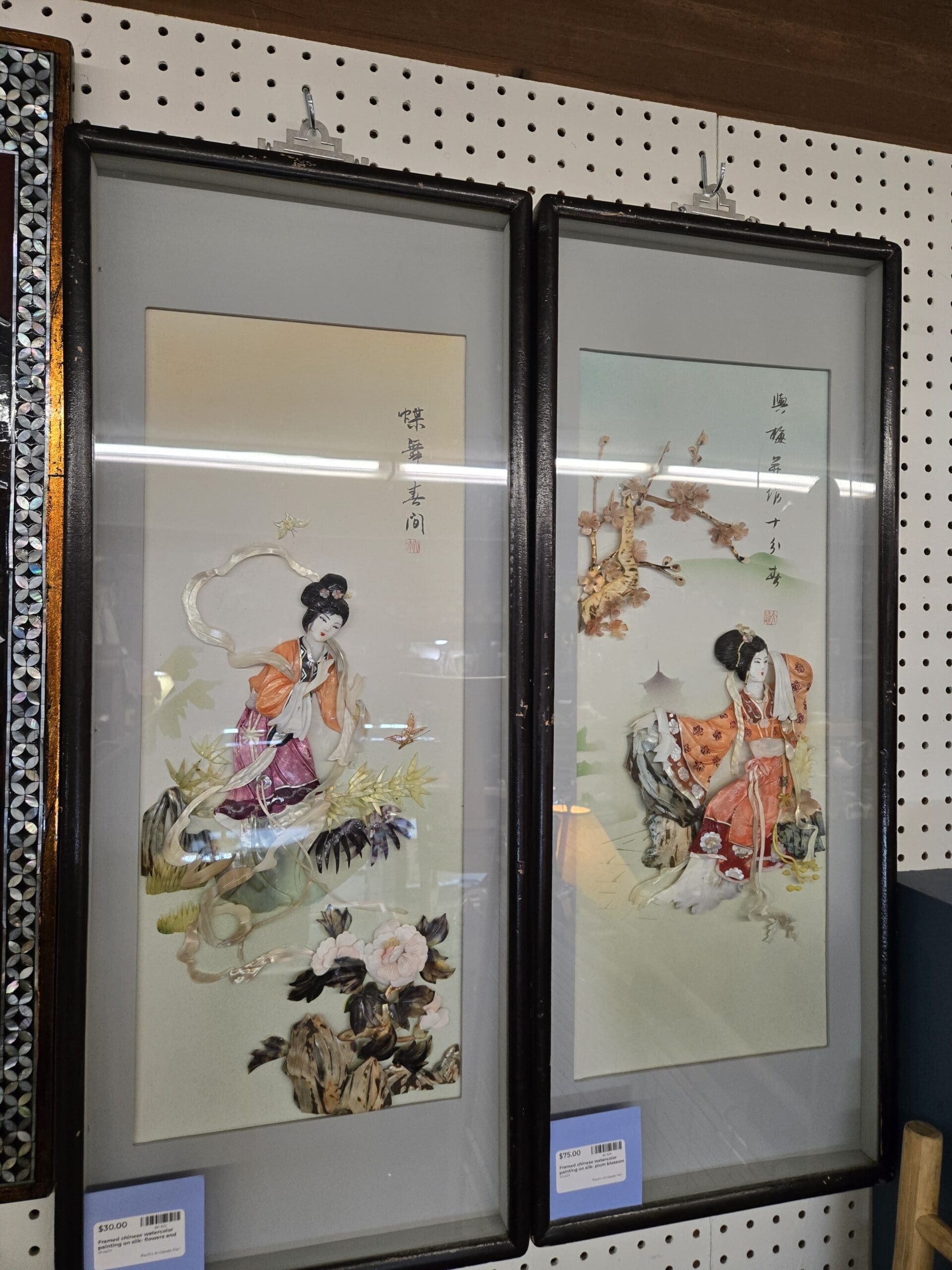Two framed Chinese decorative panels with raised relief figures of women in traditional dress on cream silk-toned grounds, the left showing a figure in orange robes with flowing scarves among butterflies and flowers, the right showing a seated figure in coral robes against a stylized pagoda landscape with plum blossoms, each panel including Chinese calligraphy and red artist seals in dark wood frames at Shop 24, Pacific Antiques Mall in Tacoma, WA.