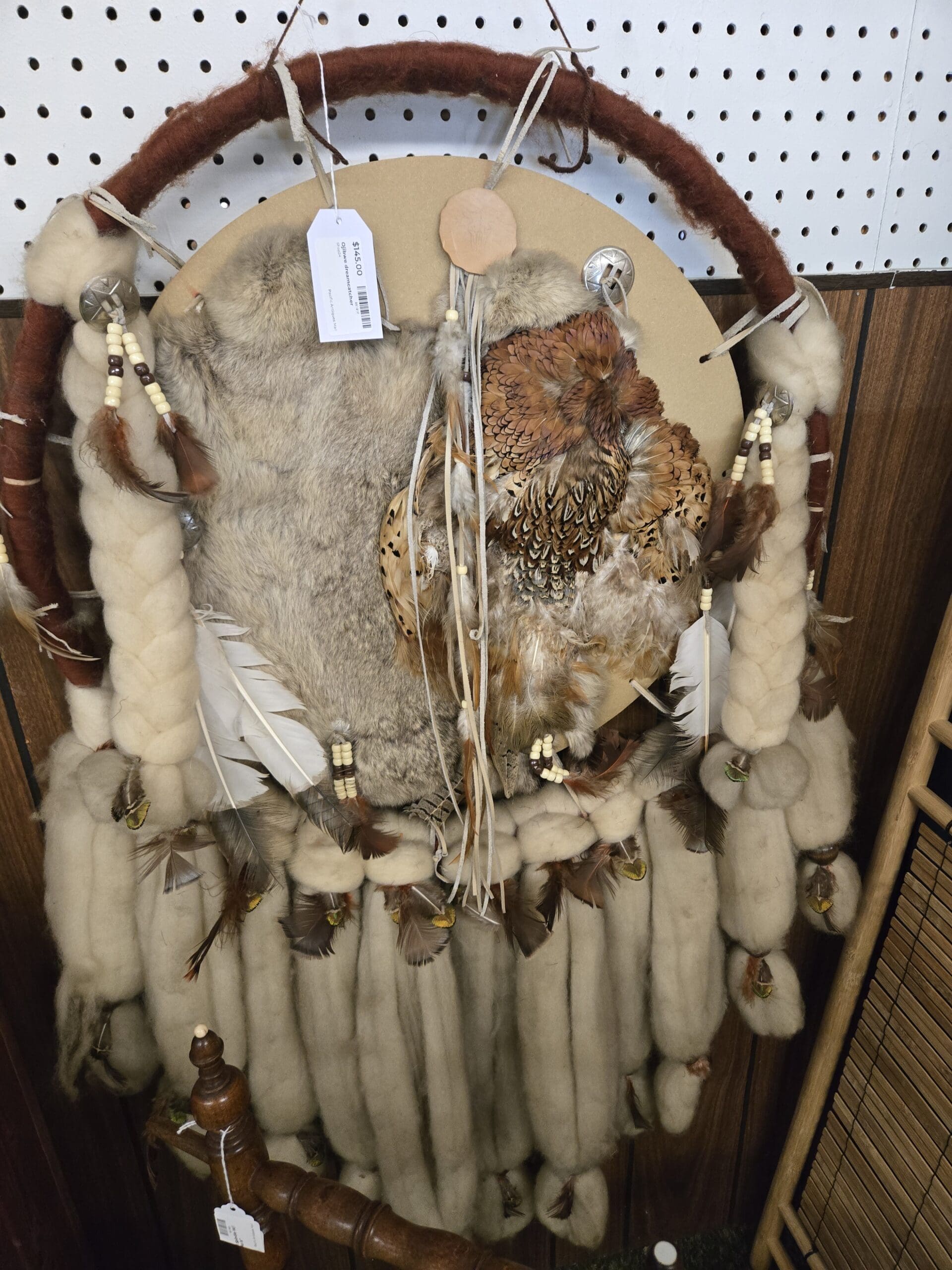 Large Plains-style dreamcatcher wall hanging on a rust-leather wrapped circular hoop, with a gray fur center, mounted brown-and-tan pheasant feather work, rows of natural cream-and-tan fur tails tipped with feathers cascading from the bottom, braided cream wool sections at the sides, beaded ties with white wood beads, and silver concho accents at Shop 24, Pacific Antiques Mall in Tacoma, WA.