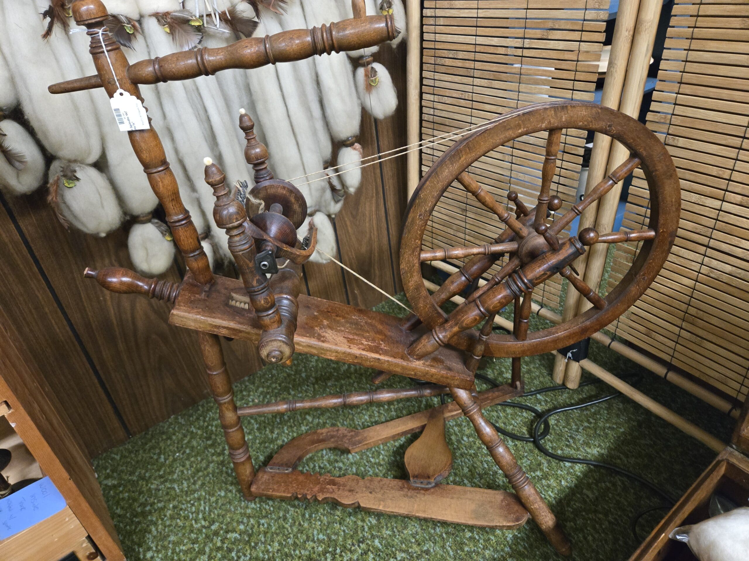 Antique wooden spinning wheel with turned spindles, large spoked drive wheel, bobbin and flyer assembly, and treadle base, set on green carpet against a bamboo screen at Shop 24, Pacific Antiques Mall in Tacoma, WA.