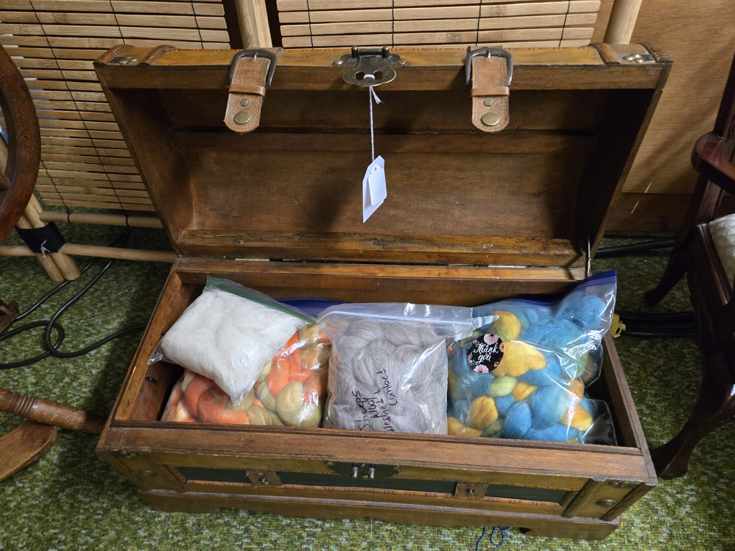 Antique flat-top wooden trunk with leather strap closures and brass hardware, opened to reveal bagged sheep wool roving in white, peach, gray combed, multicolor yellow and orange, and bright blue, with a handwritten sheeps wool combed label visible at Shop 24, Pacific Antiques Mall in Tacoma, WA.