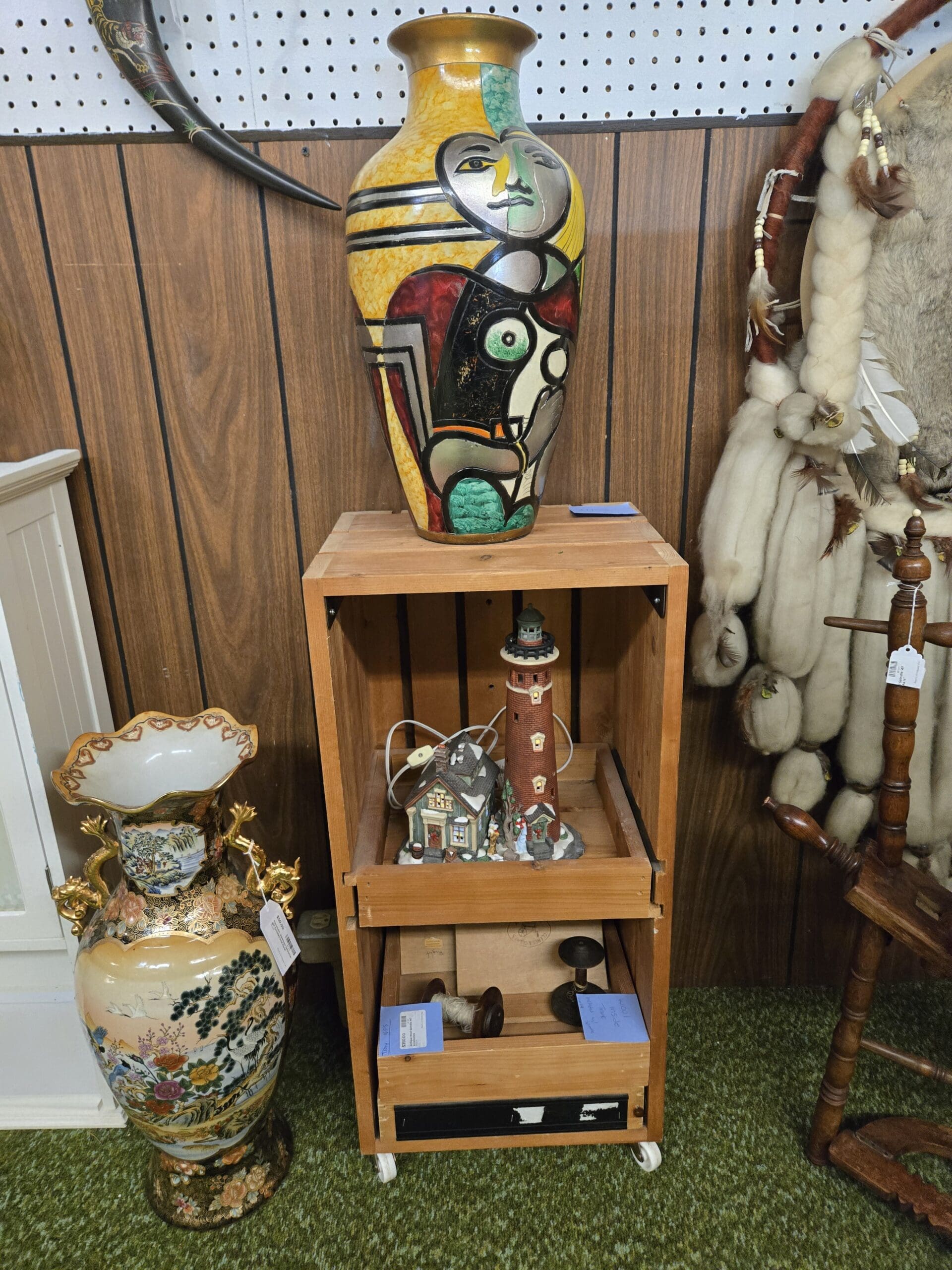 Large cubist face vase in the Picasso style with painted abstract features in mustard yellow, red, green, and black on a pine open crate shelf holding a lighted porcelain lighthouse and coastal cottage scene, beside a large Japanese Satsuma-style floor vase with hand-painted floral panels and gilded dragon-form handles on a matching plinth at Shop 24, Pacific Antiques Mall in Tacoma, WA.