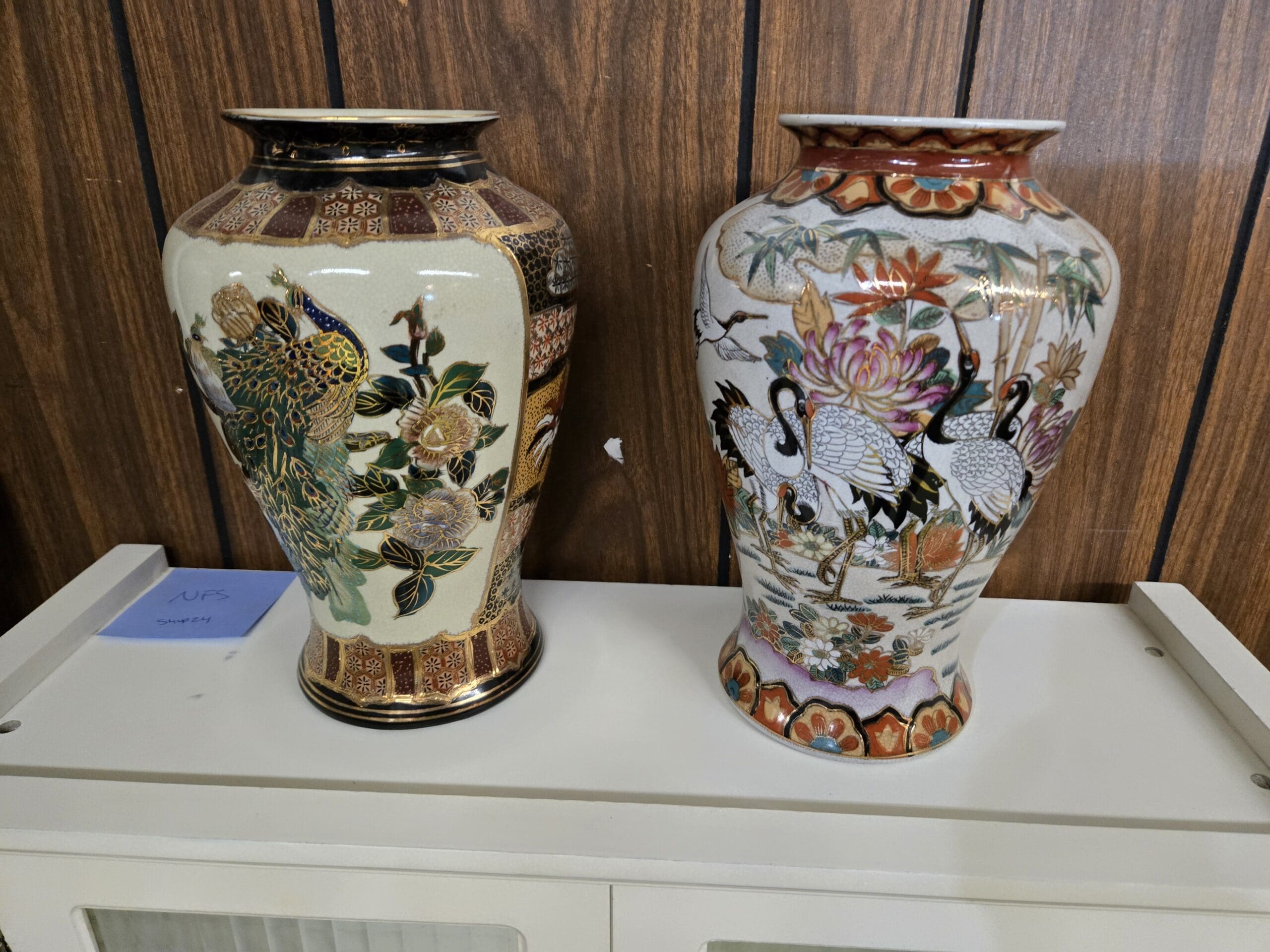 Two Japanese Satsuma-style baluster vases on a white cabinet top, the left with a cream crackle-glaze ground painted with a peacock among peonies and a cobalt blue neck with gold geometric border panels, the right with a cream crackle-glaze ground painted with red crowned cranes among bamboo and chrysanthemums framed by red lotus petal borders at Shop 24, Pacific Antiques Mall in Tacoma, WA.