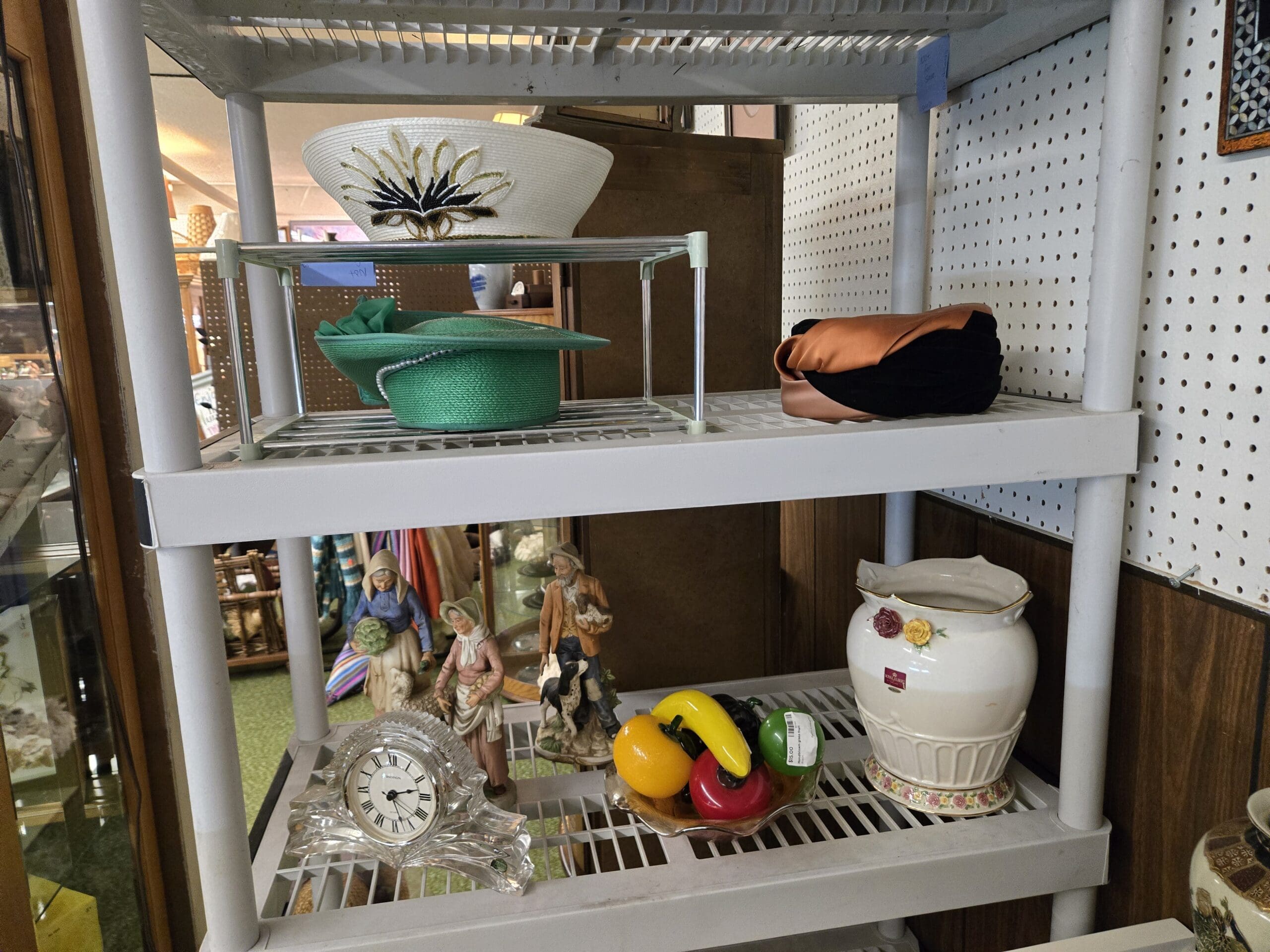 Two-tier shelving with vintage millinery on top including a white straw hat with black and gold beaded feather trim, a green straw hat with green ribbon, and a peach and black satin pillbox hat, and a lower shelf with a small lead crystal mantel clock with Roman numeral dial, two bisque figurines of an elderly couple in folk dress, a ceramic fruit display with banana, apple, pepper, and gourd in a footed bowl, and a cream English-style bone china jar with applied yellow and red roses and gilt trim at Shop 24, Pacific Antiques Mall in Tacoma, WA.