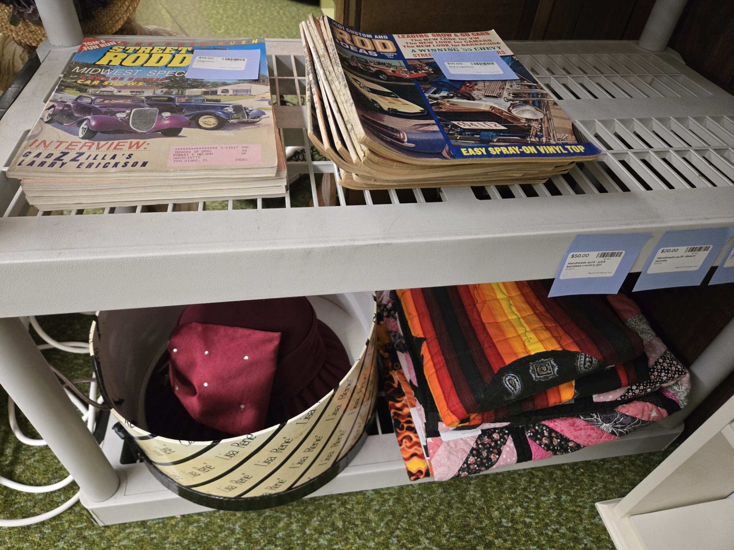 Two stacks of vintage automotive magazines including Street Rodder and Rod and Custom with hot rod and 1955 Chevy covers on a top shelf, with a Lisa Renee branded hatbox below holding a burgundy turban-style hat and a stack of handmade quilts in flame stripe, paisley, pink patchwork, and black-bordered patterns on the bottom shelf at Shop 24, Pacific Antiques Mall in Tacoma, WA.