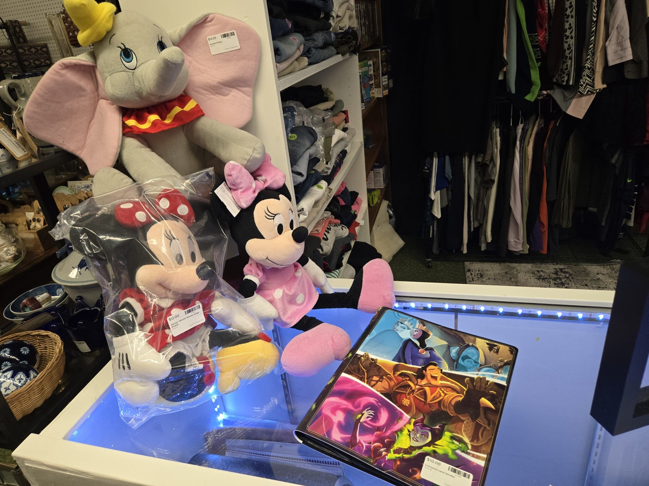A large Dumbo plush in gray with a yellow hat and red gold-star ruff, two Minnie Mouse plush toys in classic red polka-dot and pink polka-dot dresses, and a three-ring binder with Disney Villains cover art featuring Maleficent, the Evil Queen, and Ursula on a blue LED-lit counter at Shop 92, Pacific Antiques Mall in Tacoma, WA.