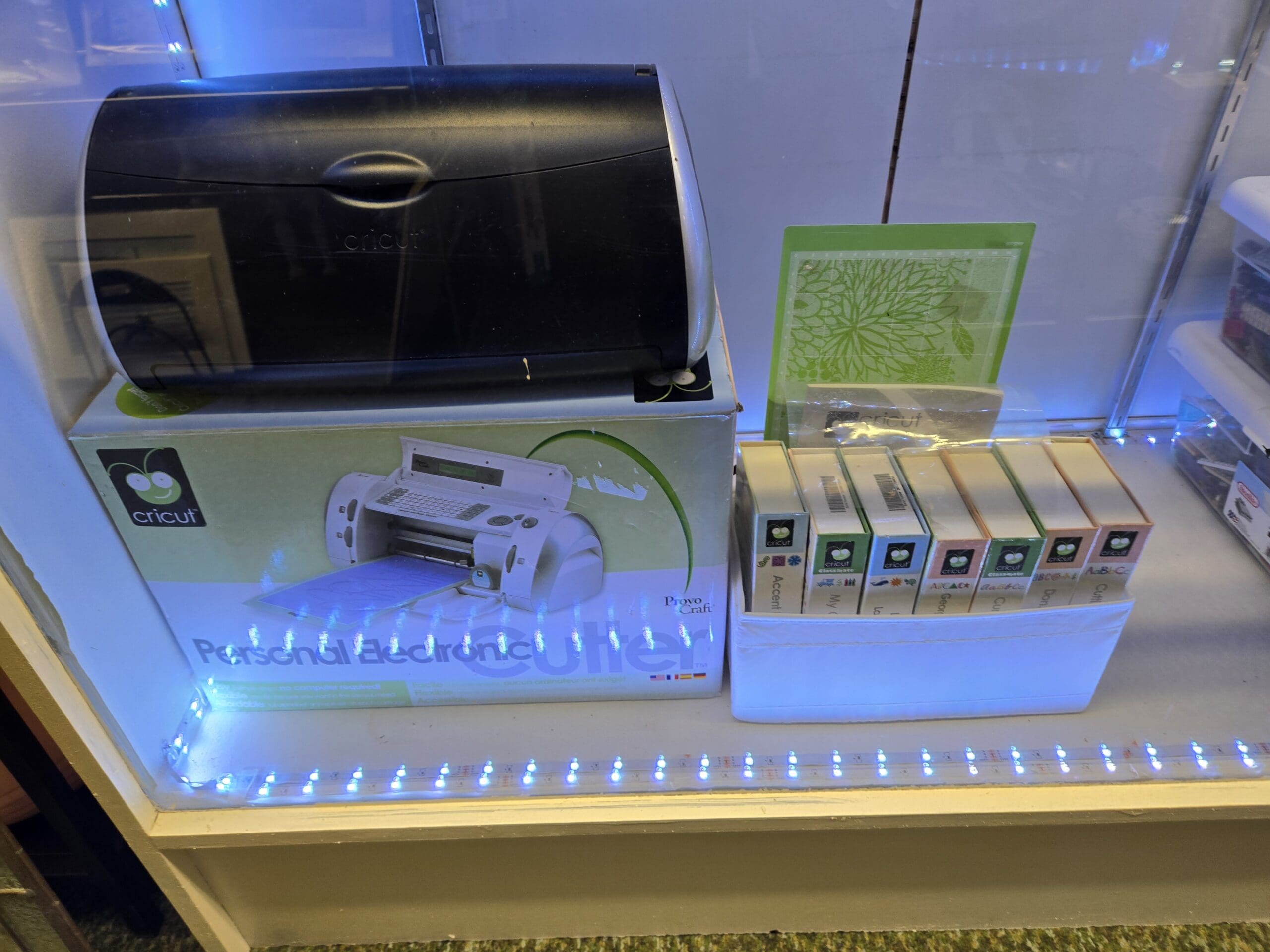 A Cricut Personal Electronic Cutter machine in black and silver by Provo Craft sitting on its original green Cricut box, with a row of seven boxed Cricut cartridges and a green Cricut die cut template inside an LED-lit display case at Shop 92, Pacific Antiques Mall in Tacoma, WA.