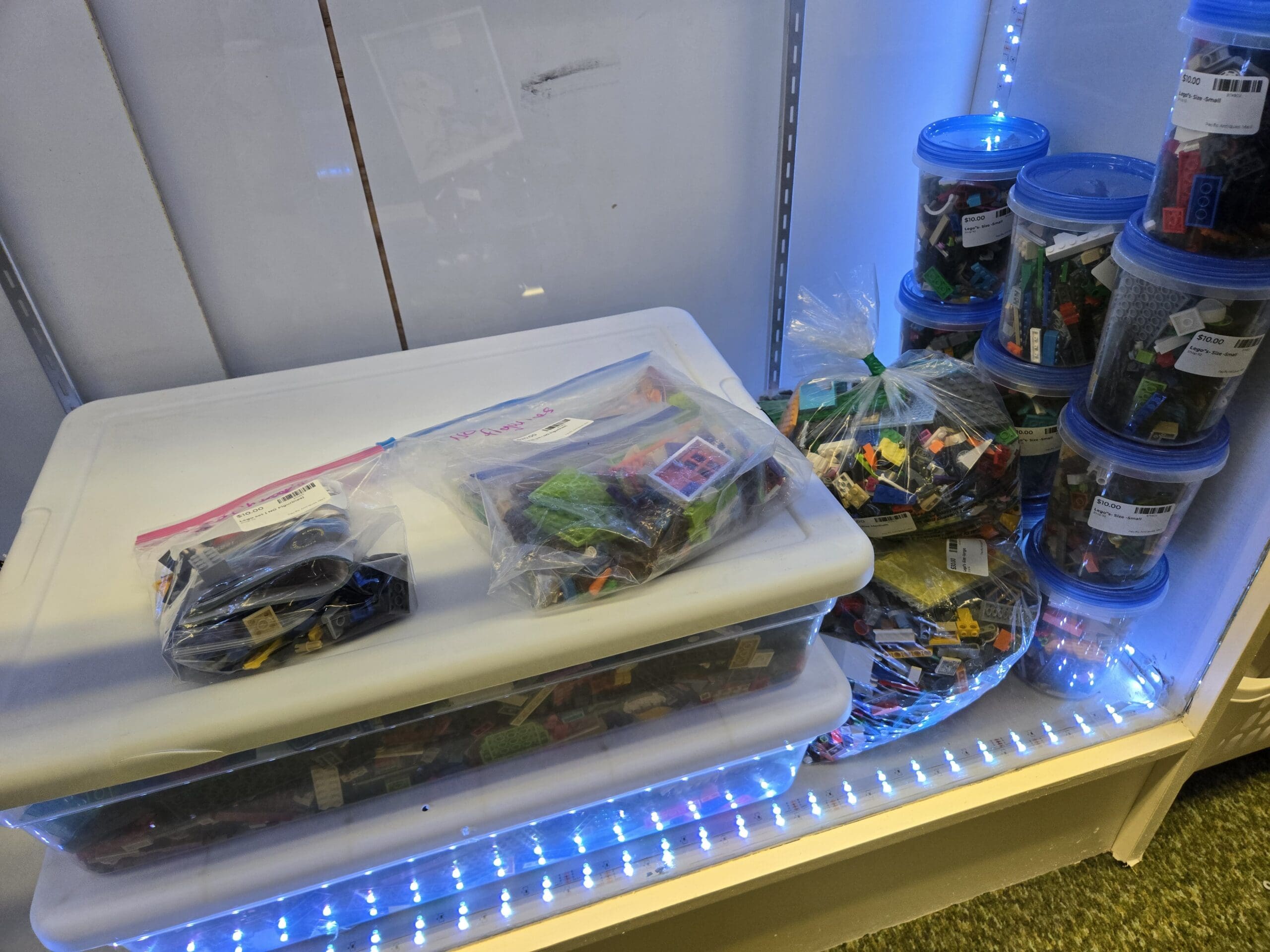 Bulk LEGO bricks in clear plastic tubs with blue lids stacked along the right side, with bagged assortments of mixed LEGO pieces on flat-topped storage bins to the left, all inside an LED-lit display case at Shop 92, Pacific Antiques Mall in Tacoma, WA.