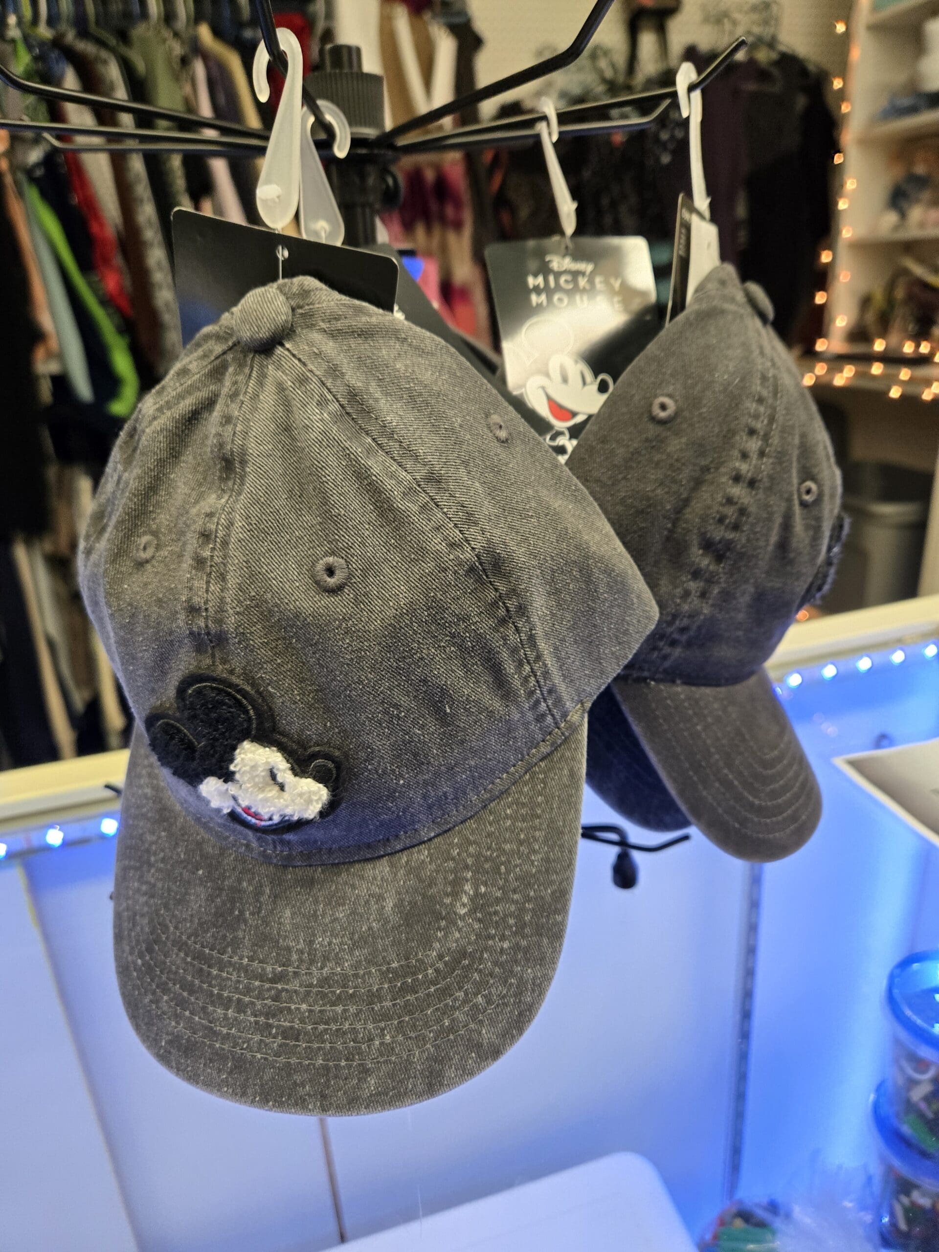 Two washed gray and navy Disney Mickey Mouse baseball caps with embroidered Mickey face appliques, hanging from a wire display rack with original Disney brand tags above the LED-lit counter at Shop 92, Pacific Antiques Mall in Tacoma, WA.