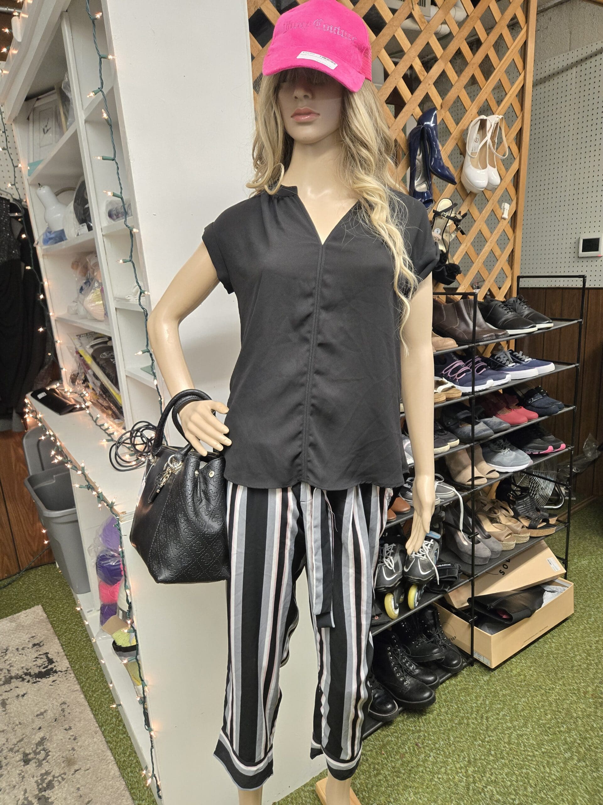 A female mannequin in a pink Juicy Couture baseball cap, blonde wig, black silky V-neck blouse, and black white and gray vertical-stripe pants, holding a black embossed-pattern handbag, posed in front of a multi-tier wire shoe rack stocked with sneakers, boots, heels, and sandals, against a wood lattice screen at Shop 92, Pacific Antiques Mall in Tacoma, WA.
