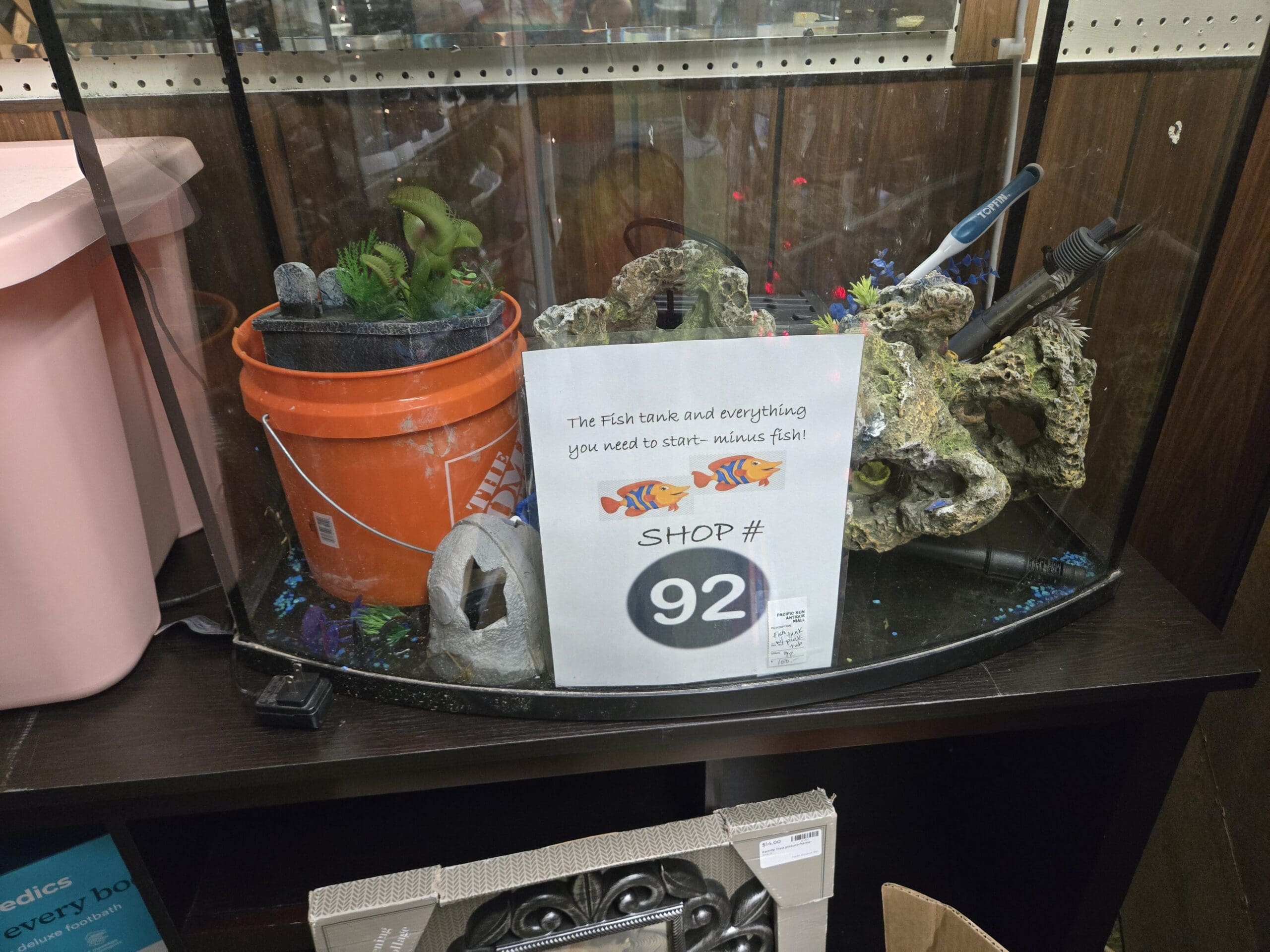 A bowfront glass aquarium tank set up with a Tetra aquarium heater, decorative rocks with cave openings, faux green plants, blue and purple gravel, a ceramic skull cave decor, a 5-gallon orange Home Depot bucket inside, and a printed sign reading the fish tank and everything you need to start, minus fish, Shop 92, at Shop 92, Pacific Antiques Mall in Tacoma, WA.