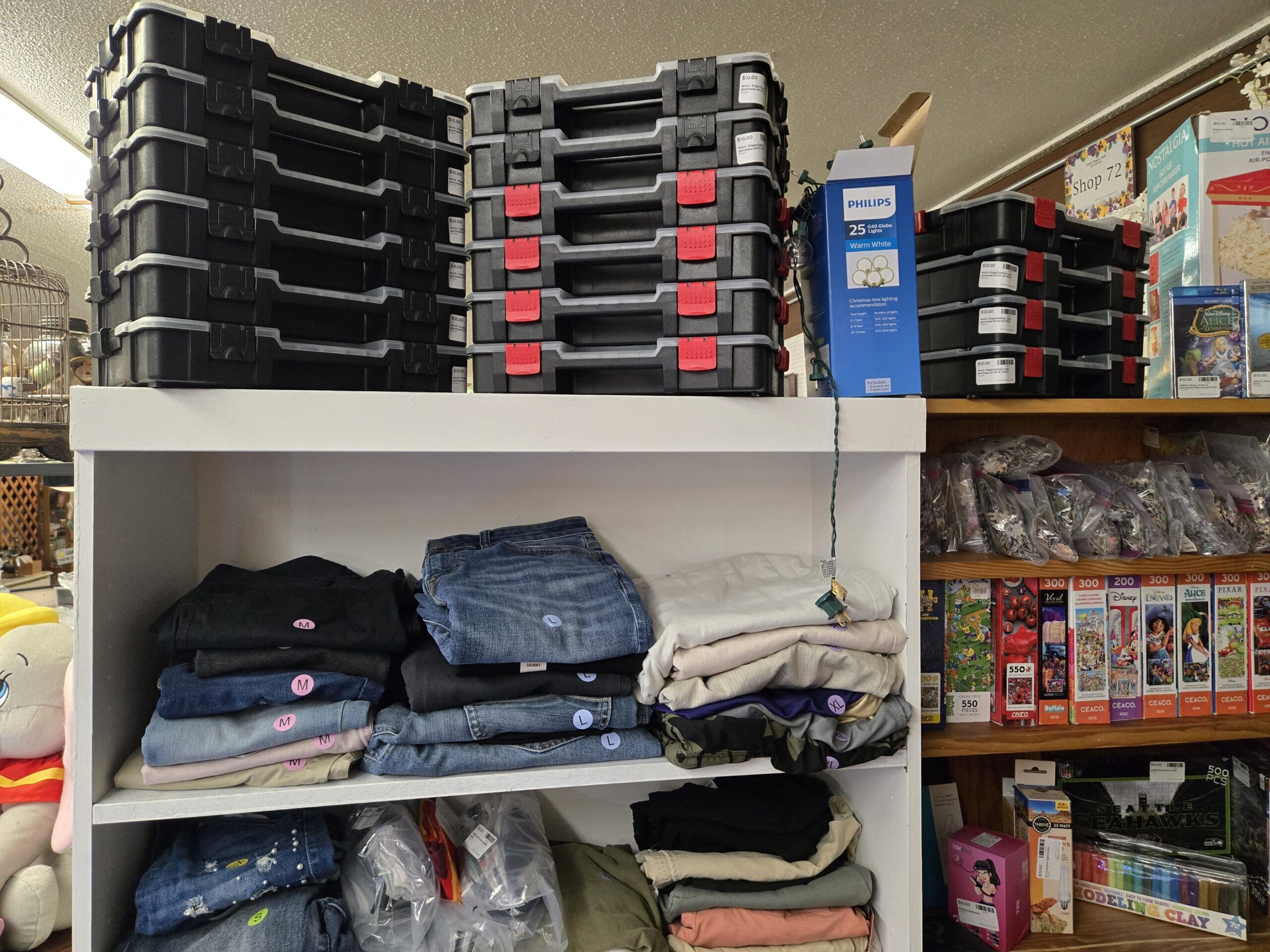 Stacks of black plastic storage organizer cases with red latches on top of a white shelving unit, beside a Philips 25 LED Globe Lights warm white box, with shelves of folded women's denim jeans, T-shirts in cream, gray, purple, and olive, bagged craft supplies, stacks of Ceaco jigsaw puzzles in Disney, Pixar, Alice, Encanto, Mickey, and Villains titles, modeling clay, and a Seattle Seahawks puzzle on the wood shelving to the right at Shop 92, Pacific Antiques Mall in Tacoma, WA.