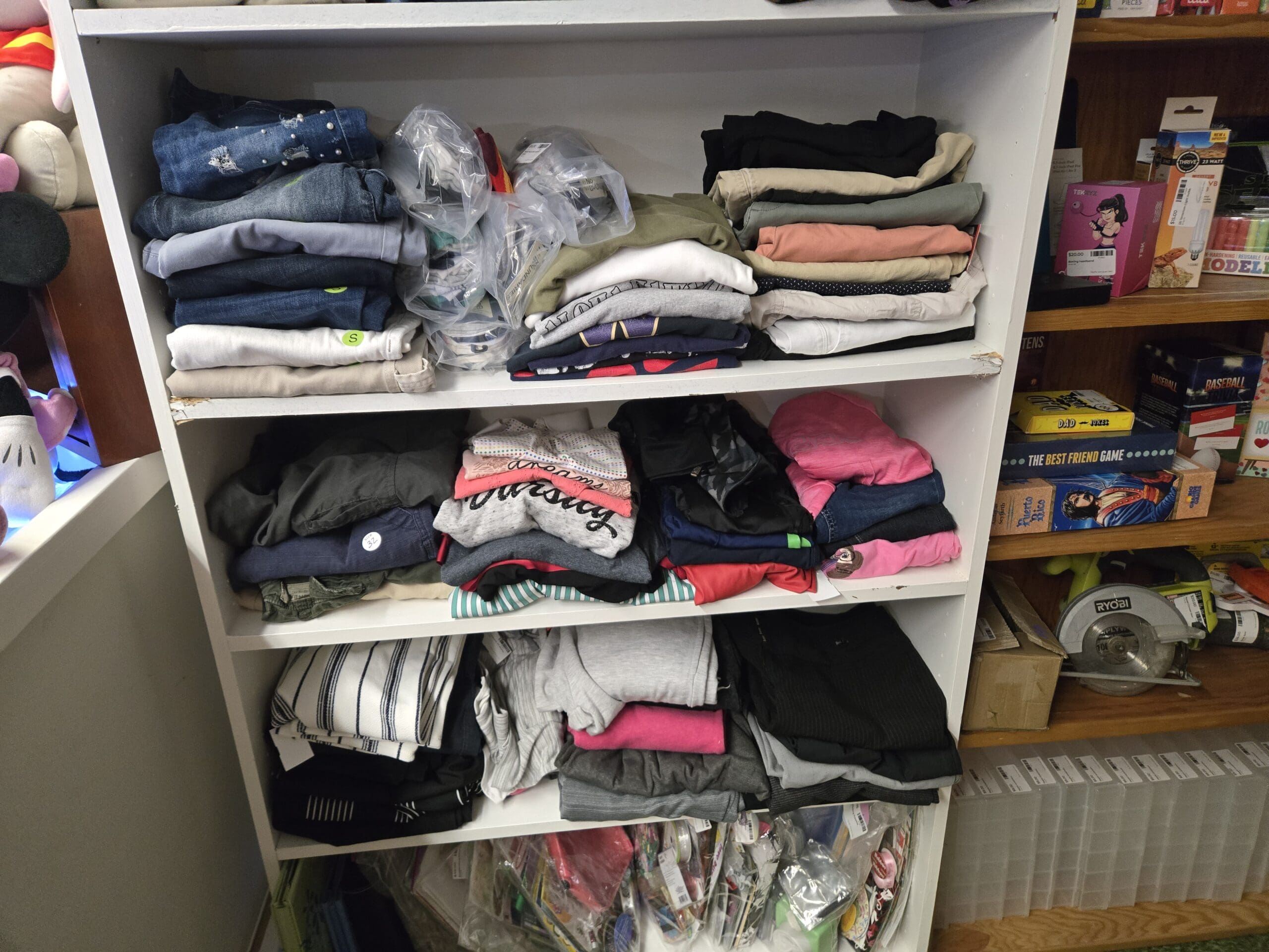 A white shelving unit stacked with folded women's clothing including jeans, varsity sweatshirts, knit tops in pink, gray, red, and navy, and striped pants, with a wood shelving unit to the right holding board and card games including The Best Friend Game, Puerto Rico, Baseball Trivia, and Dad Jokes, modeling clay, and a yellow Ryobi cordless circular saw on the lower shelf at Shop 92, Pacific Antiques Mall in Tacoma, WA.