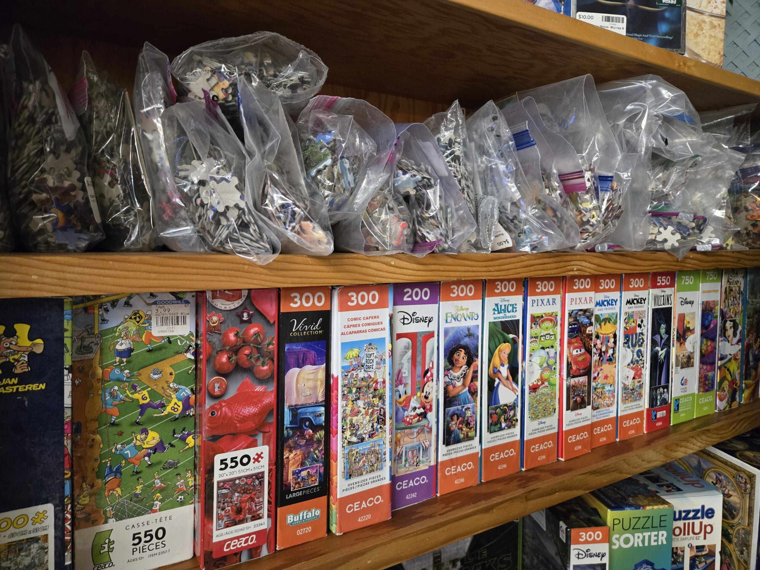 A puzzle shelf with bagged loose jigsaw puzzle pieces on the upper level and a long row of boxed jigsaw puzzles below ranging from 200 to 750 pieces by Ceaco and Buffalo Games, including Disney Encanto, Alice in Wonderland, Pixar, Mickey, Mickey and Friends, and Villains titles, a Vivid Collection tomato puzzle, a Comic Capers 300-piece, a Casse-tete 550-piece football puzzle, and a Disney Puzzle RollUp at Shop 92, Pacific Antiques Mall in Tacoma, WA.