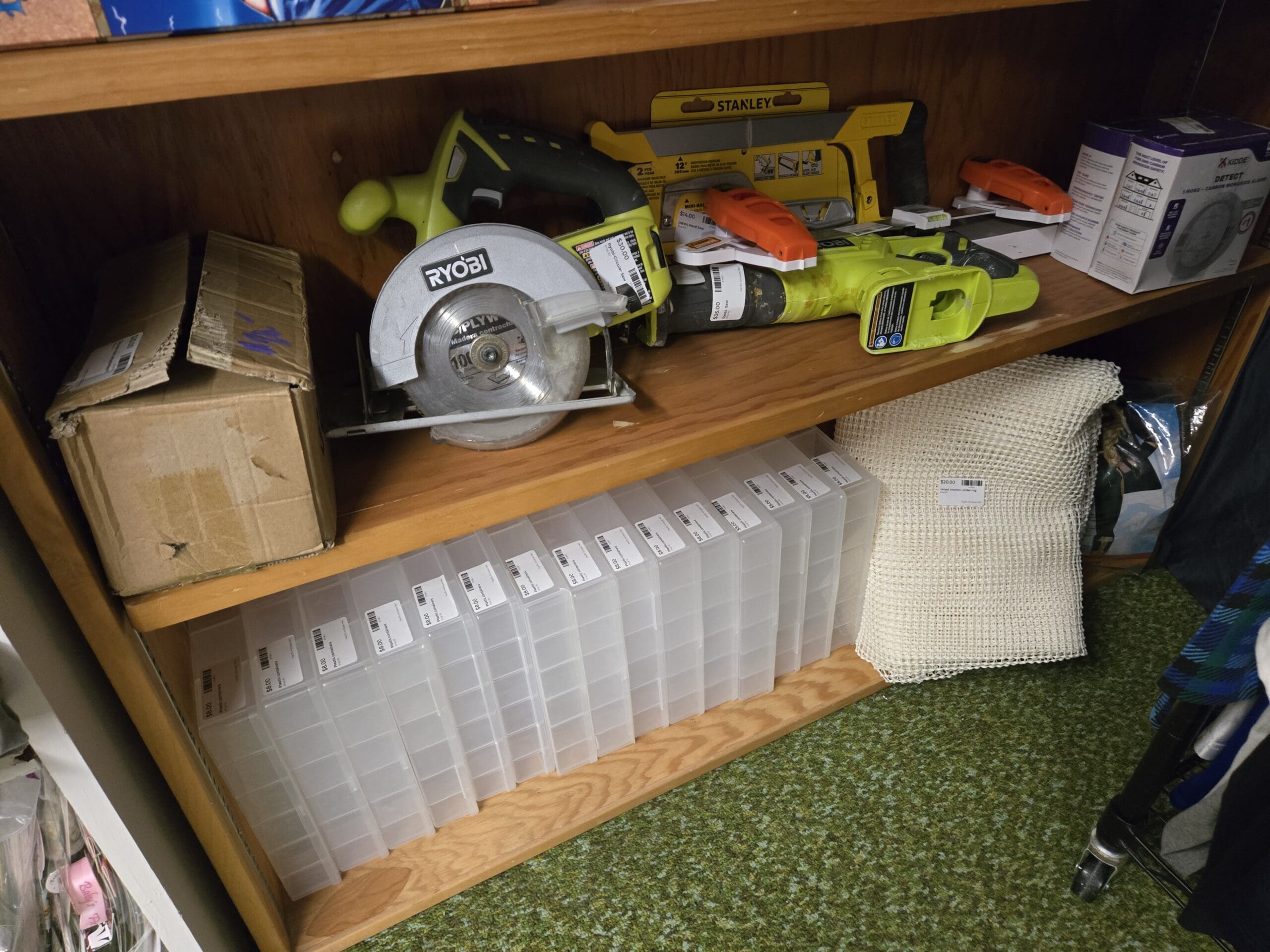 A wood shelving unit with a Ryobi cordless circular saw, a packaged Stanley hand saw, additional Ryobi cordless tools, and a Kidde smoke and carbon monoxide detector box on the upper shelf, a row of clear plastic compartmented organizer cases on the lower shelf, a sealed cardboard box, and a folded cream open-knit blanket at Shop 92, Pacific Antiques Mall in Tacoma, WA.
