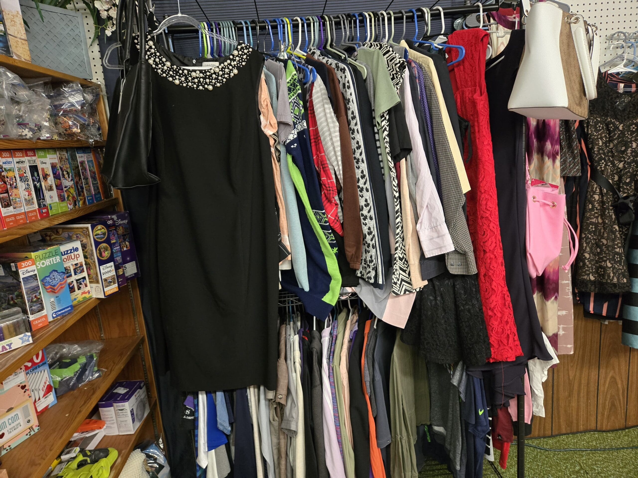 A packed clothing rack with a black sleeveless cocktail dress with pearl-beaded neckline in front, dresses and blouses behind it in peach, green, black-and-white print, lavender, red lace, pink, and tie-dye, two handbags hanging above in black faux leather and white-and-brown, with a wood shelf to the left stocked with Ceaco and Buffalo Games Disney and Mickey jigsaw puzzles, boxed sandals, and bagged craft supplies at Shop 92, Pacific Antiques Mall in Tacoma, WA.