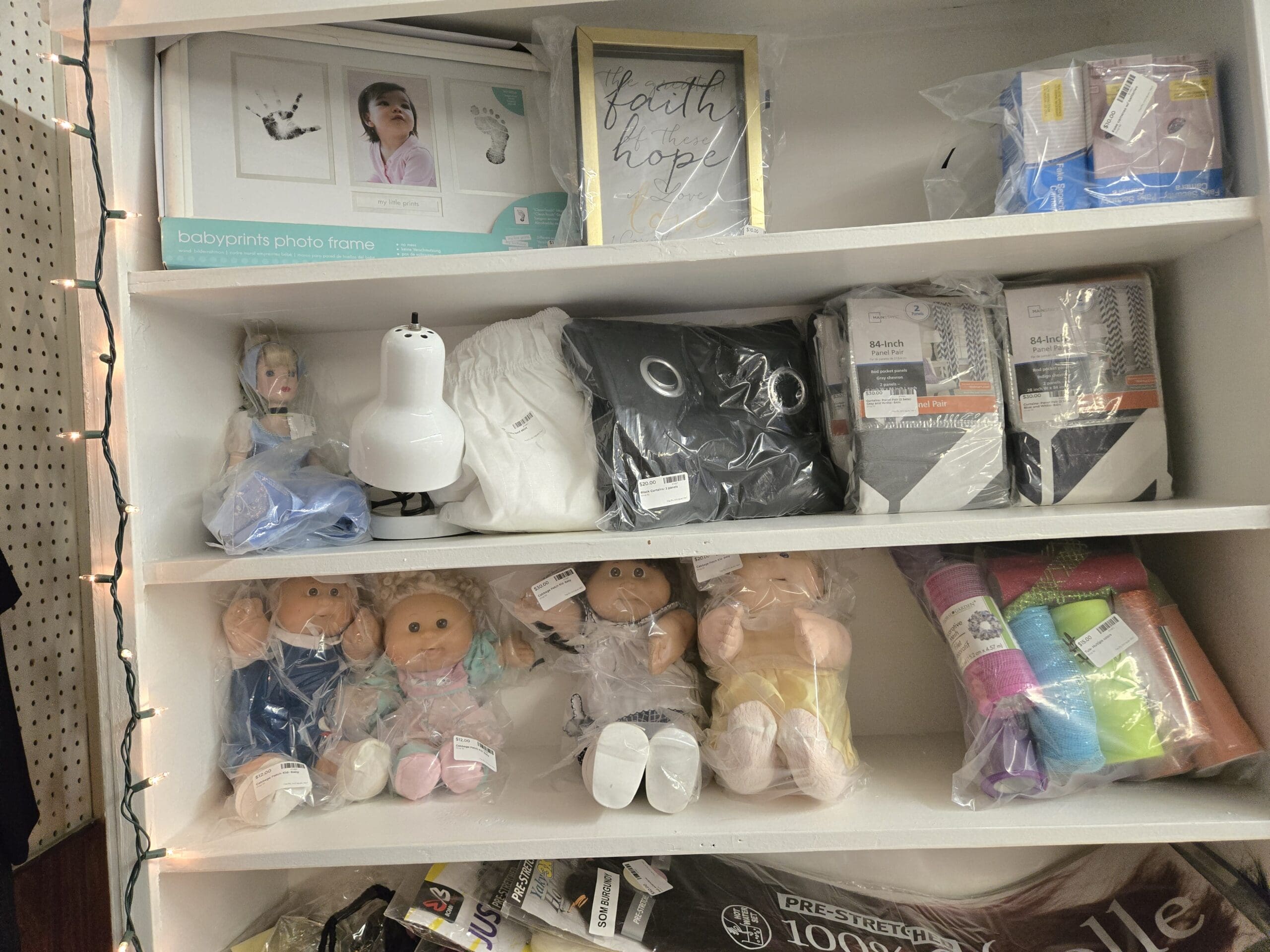 A white shelving unit with a Babyprints handprint and footprint photo frame and a gold-trim faith hope love framed wall sign on the top shelf, a Disney Cinderella doll in a blue gown and tiara beside a small white space heater and packaged Mainstays 84-inch gray and white curtain panels on the middle shelf, and four Cabbage Patch Kids dolls sealed in cellophane bags with bundles of pastel mesh decorative ribbon on the lower shelf at Shop 92, Pacific Antiques Mall in Tacoma, WA.
