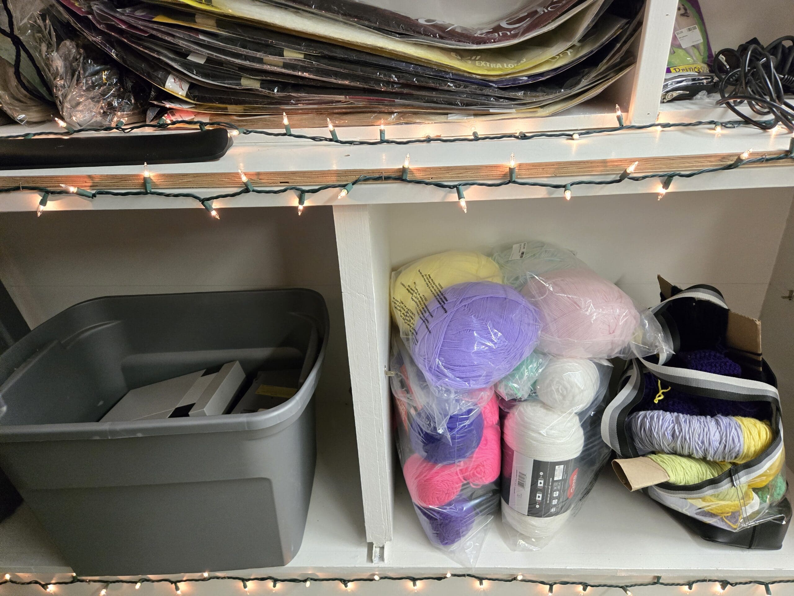 Upper shelf stacked with packages of Afrelle Kanekalon pre-stretched braiding hair extensions in multiple colors, and a lower shelf with a gray plastic storage tote of files beside bagged skeins of yarn in yellow, lavender, hot pink, mint green, white, and magenta, plus a black-and-white striped tote holding crocheted yarn lengths in yellow, lilac, and purple at Shop 92, Pacific Antiques Mall in Tacoma, WA.