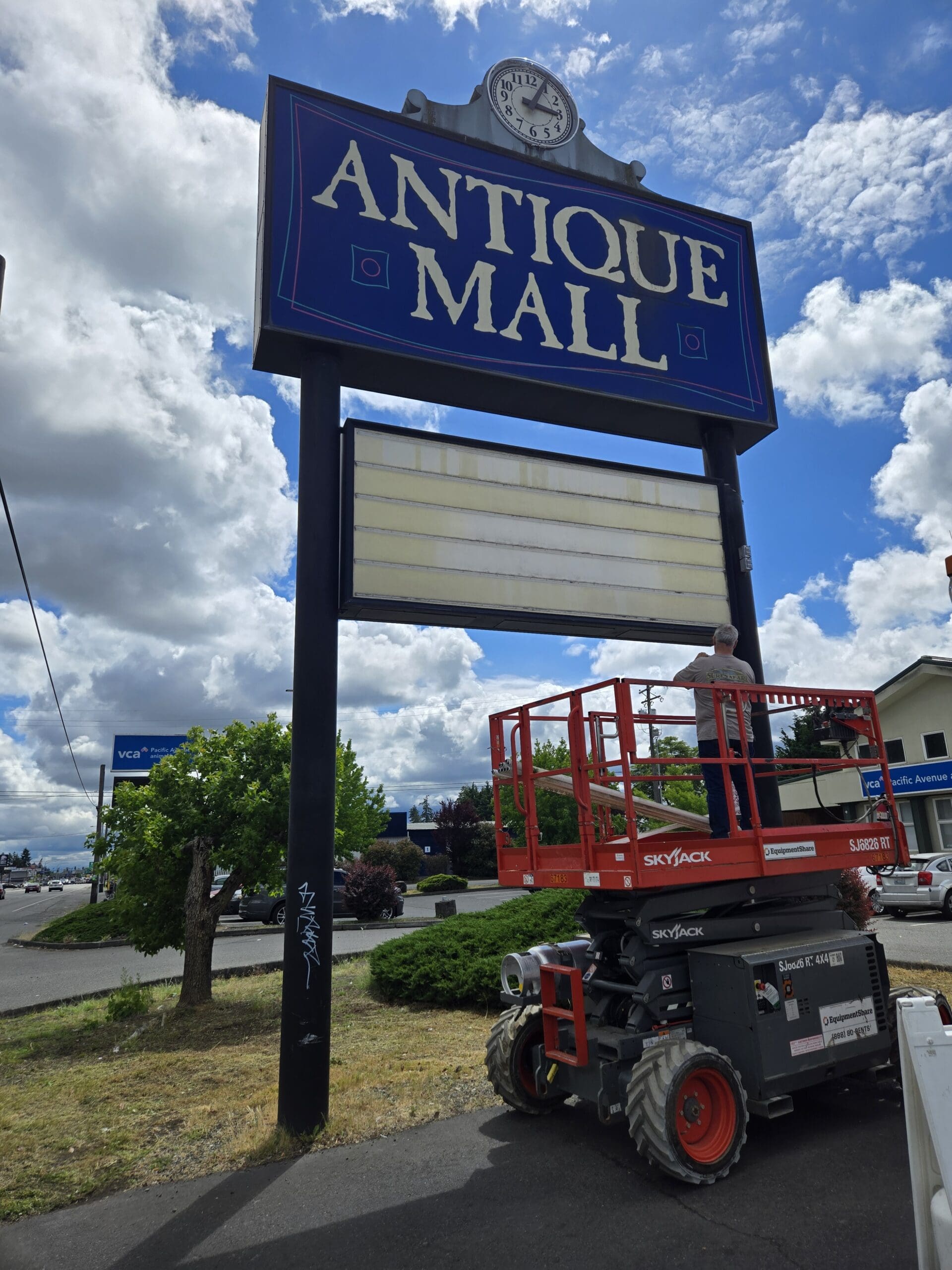 The vintage Pacific Antiques Mall pylon sign on Pacific Avenue with the deep blue ANTIQUE MALL panel and Arabic-numeral clock at top, an empty white letter-board marquee below, and a SkyJack scissor lift from Equipment Share positioned alongside with a sign technician on the platform during service work at Pacific Antiques Mall in Tacoma, WA.