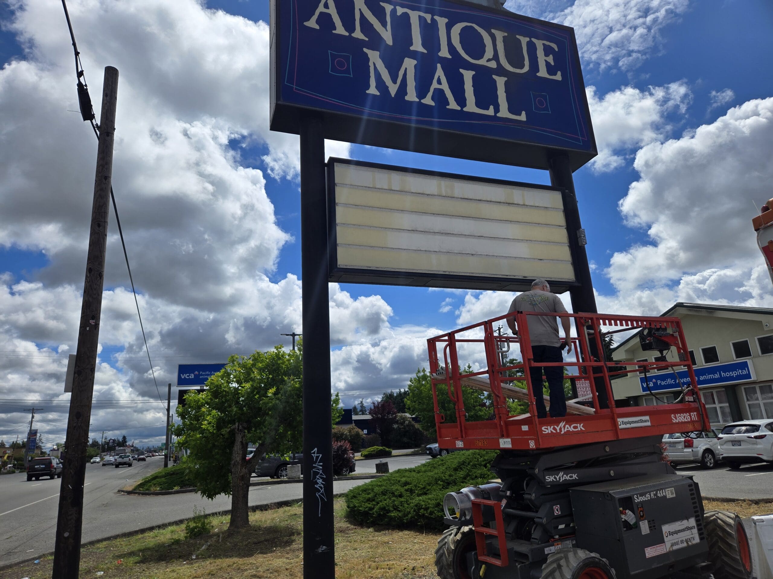 Closer view of the Pacific Antiques Mall pylon sign during service work, showing the painted blue ANTIQUE MALL panel, the empty white letter-board marquee below, and a sign technician on the orange SkyJack scissor lift platform extended to sign height, with Pacific Avenue traffic and the VCA Pacific Avenue Animal Hospital visible behind at Pacific Antiques Mall in Tacoma, WA.