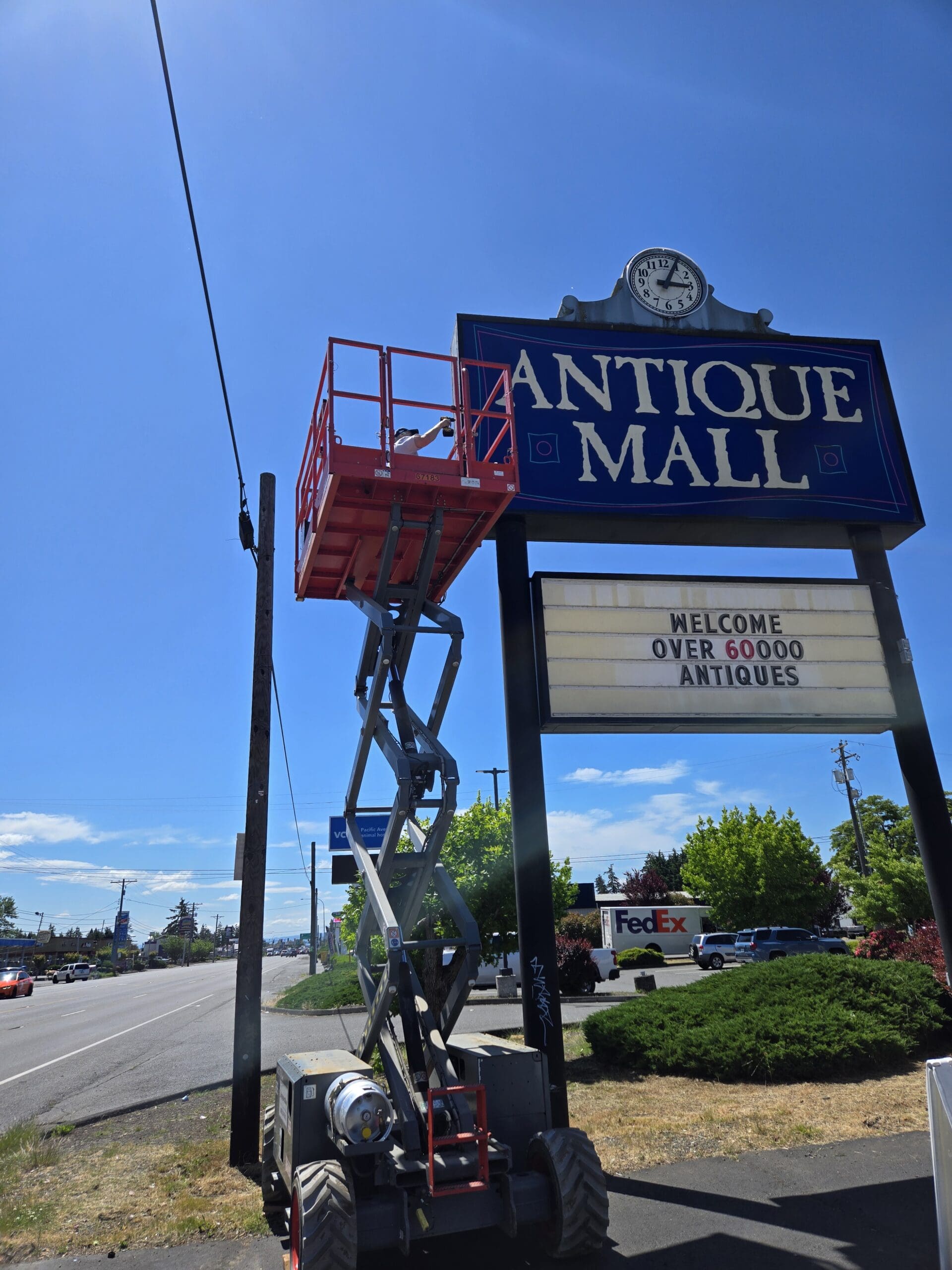 The Pacific Antiques Mall pylon sign with the deep blue ANTIQUE MALL panel and clock at top and a refreshed letter-board marquee reading WELCOME OVER 60000 ANTIQUES below, with the SkyJack scissor lift fully extended in the foreground and a FedEx delivery truck passing on Pacific Avenue behind at Pacific Antiques Mall in Tacoma, WA.
