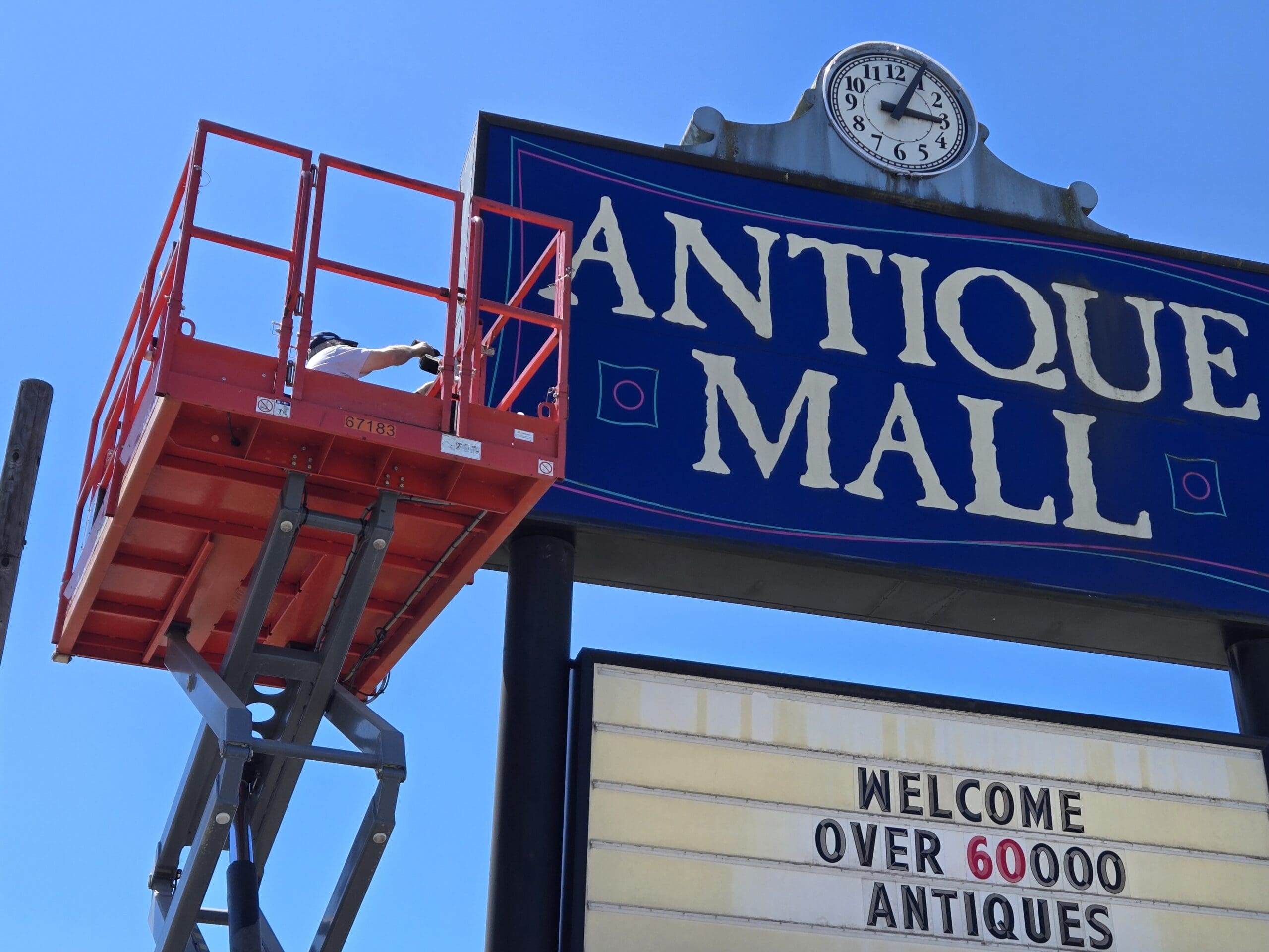Close upward view of the Pacific Antiques Mall pylon sign with the Arabic-numeral clock and the ANTIQUE MALL panel painted in white and pink-coral lettering on deep blue, with a sign technician on the orange SkyJack platform reaching toward the right edge of the sign at Pacific Antiques Mall in Tacoma, WA.